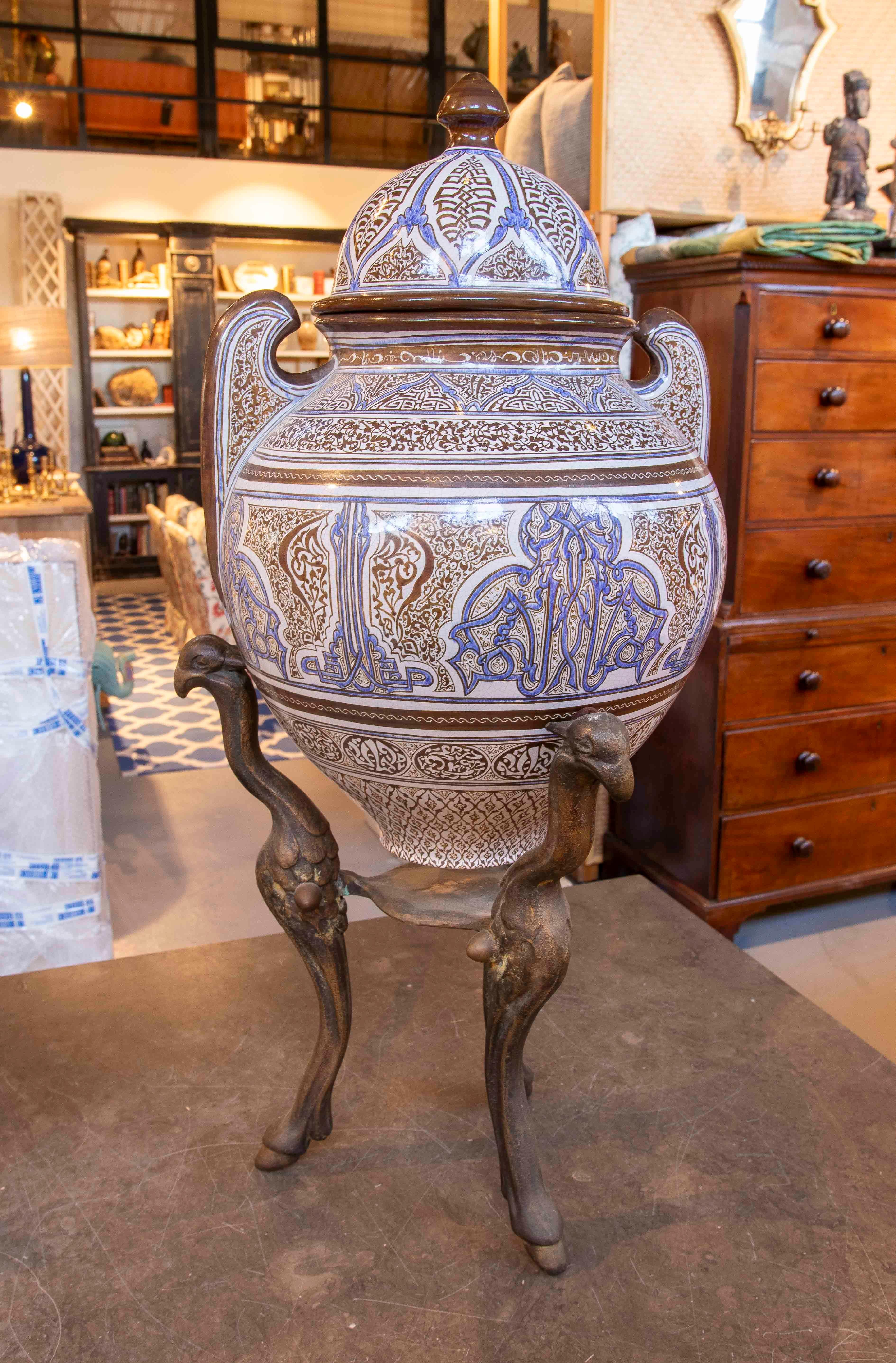 Impressive Arabic-inspired jar made of glazed ceramic with rich calligraphic and geometric motifs in cobalt blue and brown on a white background. The piece is topped with an ornate lid and rests on an elegant wrought iron tripod with zoomorphic