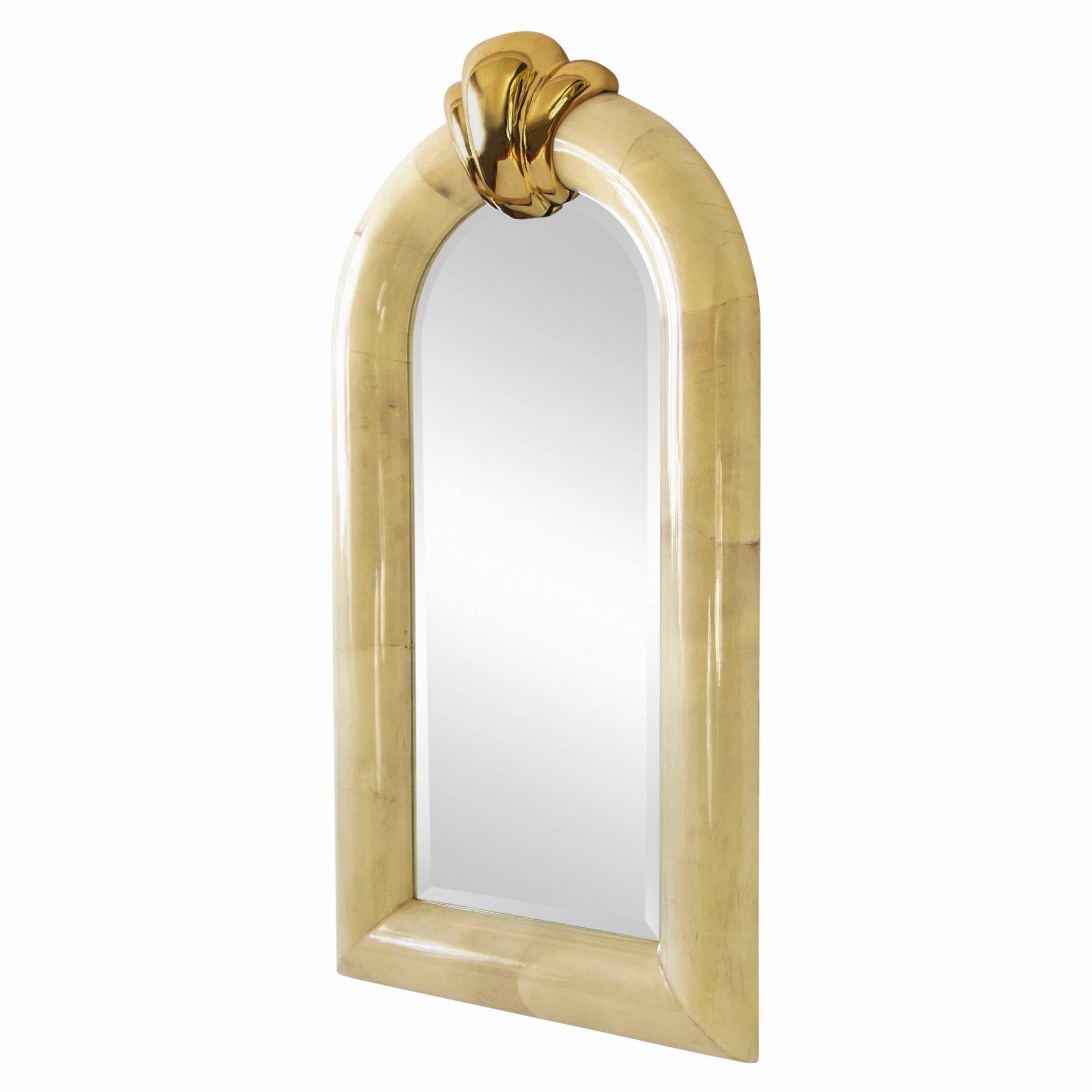 1980s Karl Springer inspired goatskin beveled mirror with a large solid brass keystone crest. The frame is made of high-gloss lacquered cream-color untanned goat skin or parchment stretched over a 5