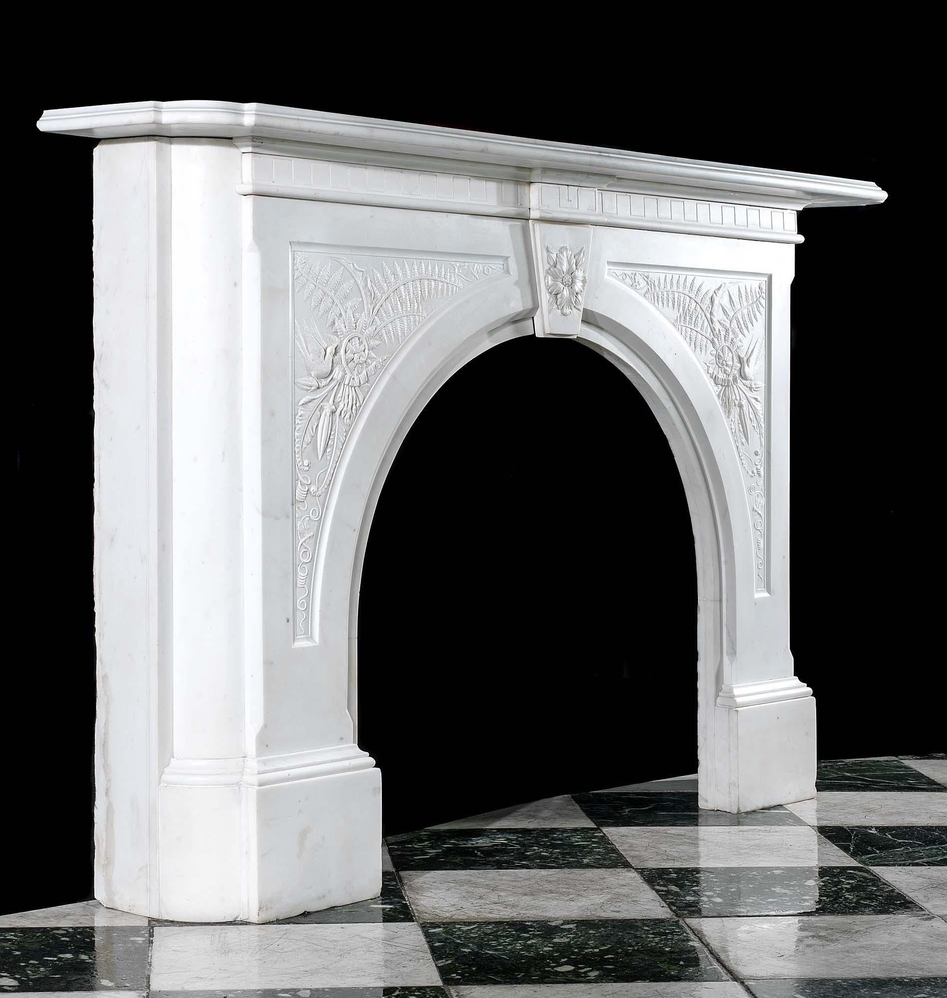 A large arched Victorian chimneypiece in pure white Statuary Marble. The wide moulded shelf rests above an arched opening set with a central floral keystone flanked by carved spandrels of birds perched on stylised fern sprays with descending buds