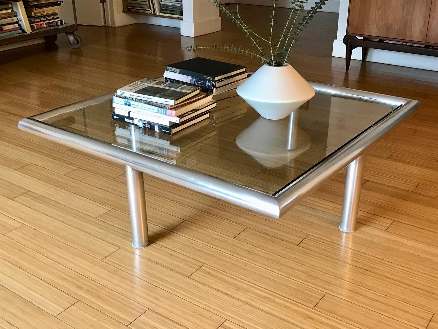 Simple and clever architectural design with a subtle unorthodox aesthetic. 
Legs positioned in the middle of the frame.
Tubular brushed aluminum (minor wear, patina).
New glass top. 
Great for any interior.
  