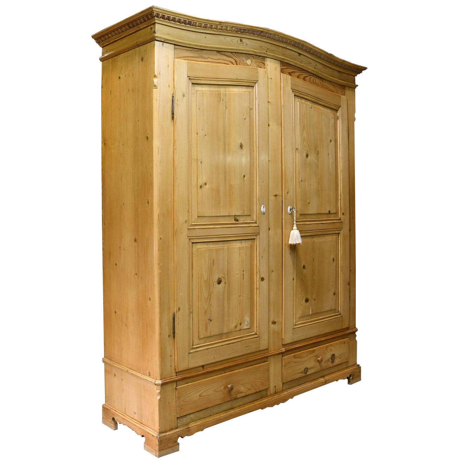 A beautiful armoire in light-colored pine with arched bonnet embellished with a carved cornice, two arched doors on outside hinges, with two raised panels on each door, and a center divide between them. Armoire has two bottom drawers and rests on a