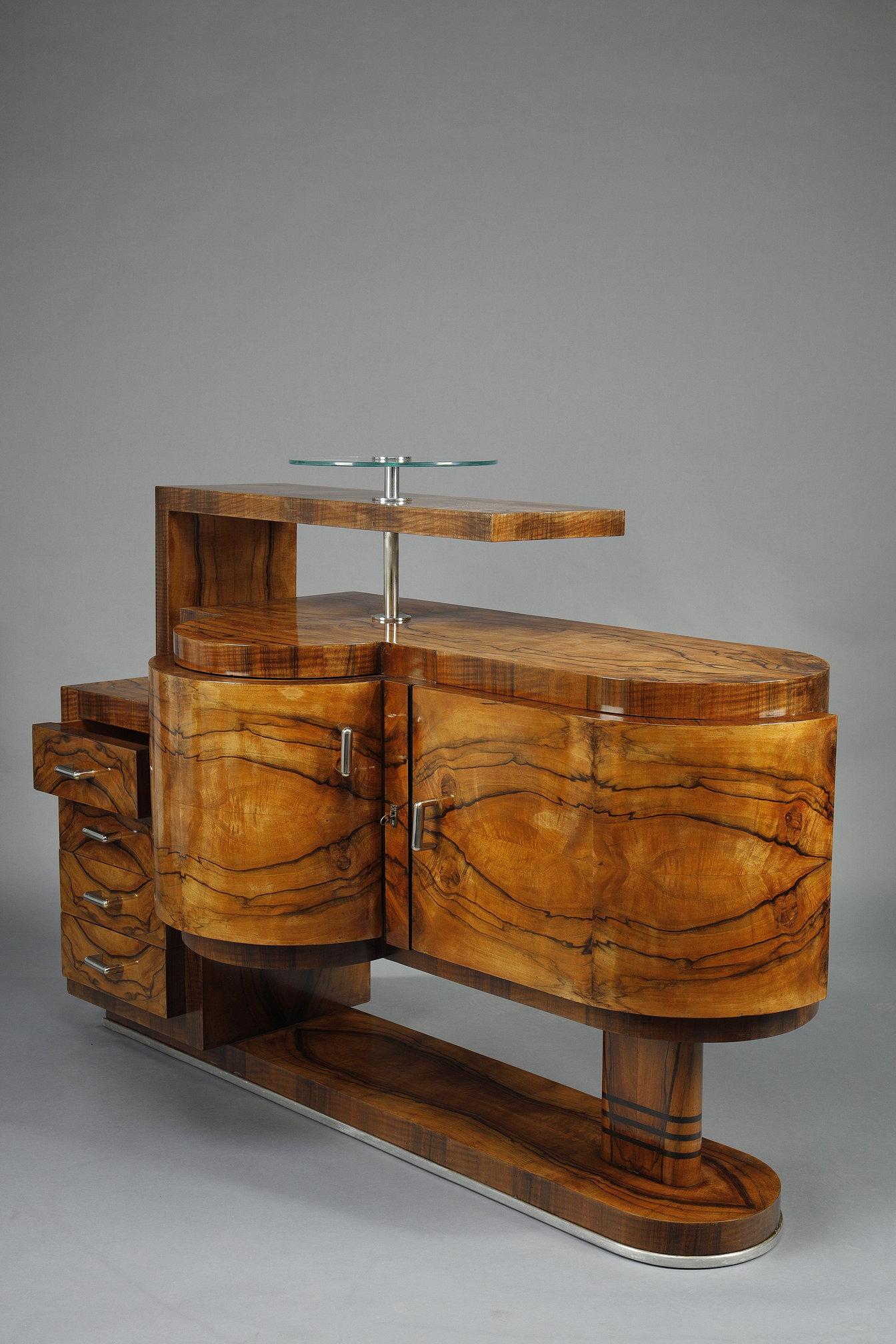 Meuble bar d'époque Art Déco en noyer et placage de noyer ouvrant à deux portes et quatre tiroirs. Base et poignées chromées. Un plateau circulaire en verre. Sa forme architecturale évoque les formes et les lignes modernistes de l'époque.

Un autre