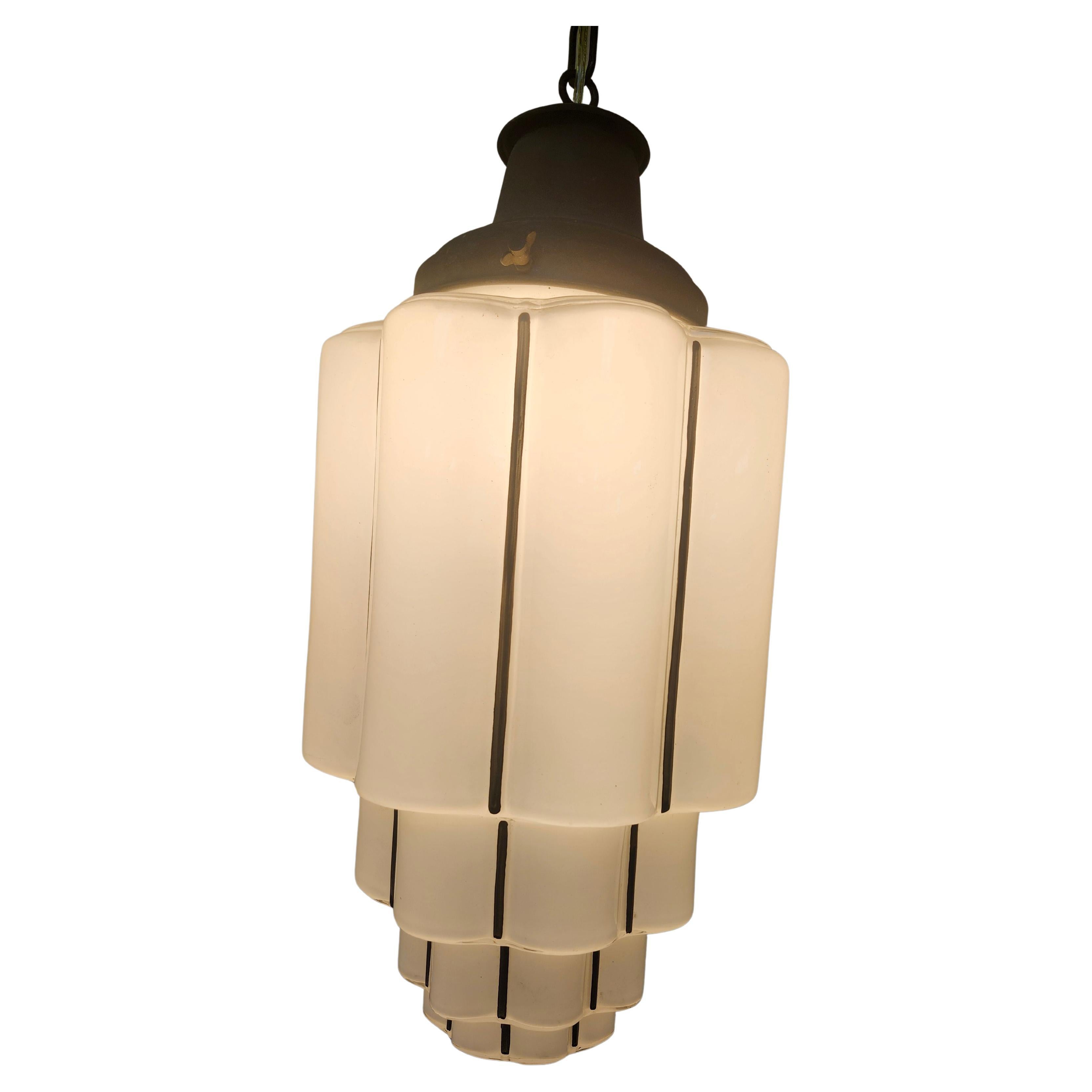 Sencilla pero elegante lámpara colgante Art Decó en forma de rascacielos de cristal de leche con bordes redondeados. Recableado con la toma de porcelana original en excelente estado. Incluye tejadillo en forma de cúpula.