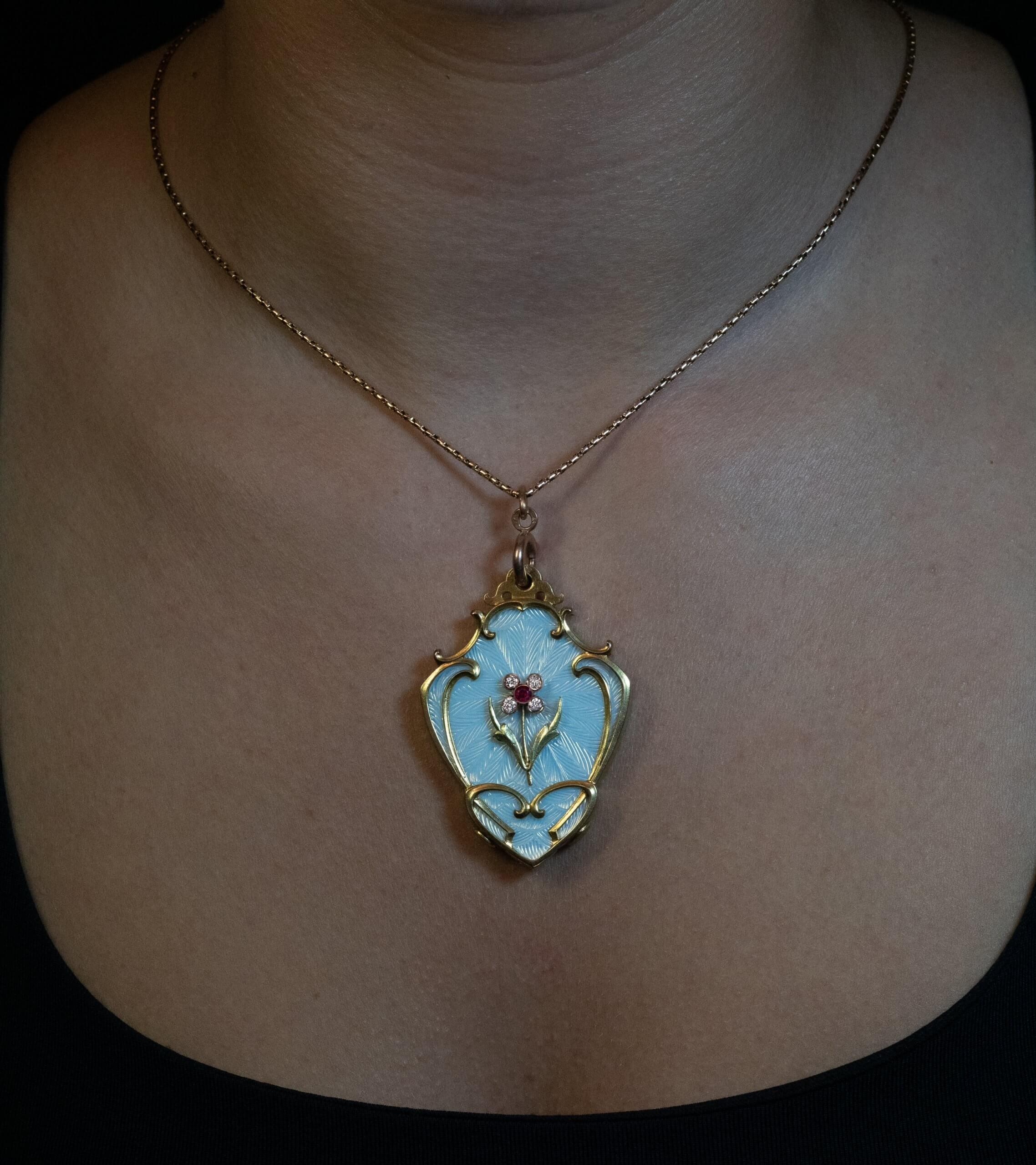 Circa 1910  
Questo grande ciondolo ornato in oro 14K è stato progettato secondo il gusto Art Nouveau dell'epoca. Il ciondolo è impreziosito da un eccellente smalto guilloché bianco-bluastro. Al centro è applicato un fiore di rubino e diamante. 