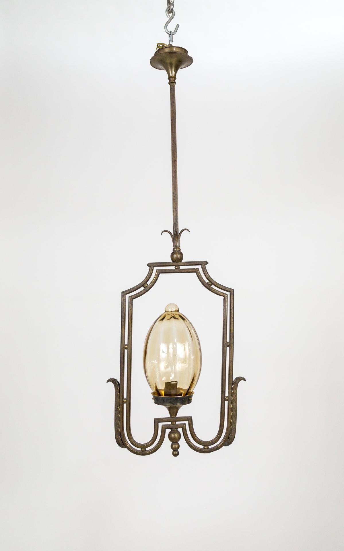 A large pendant light with a dark, patinated metal structure framing an egg-shaped glass shade. The glass has a subtle brown tint and ribbed texture. The fixture is a stylized, rectangular shape, decorated with sphere and leaf accents and a long