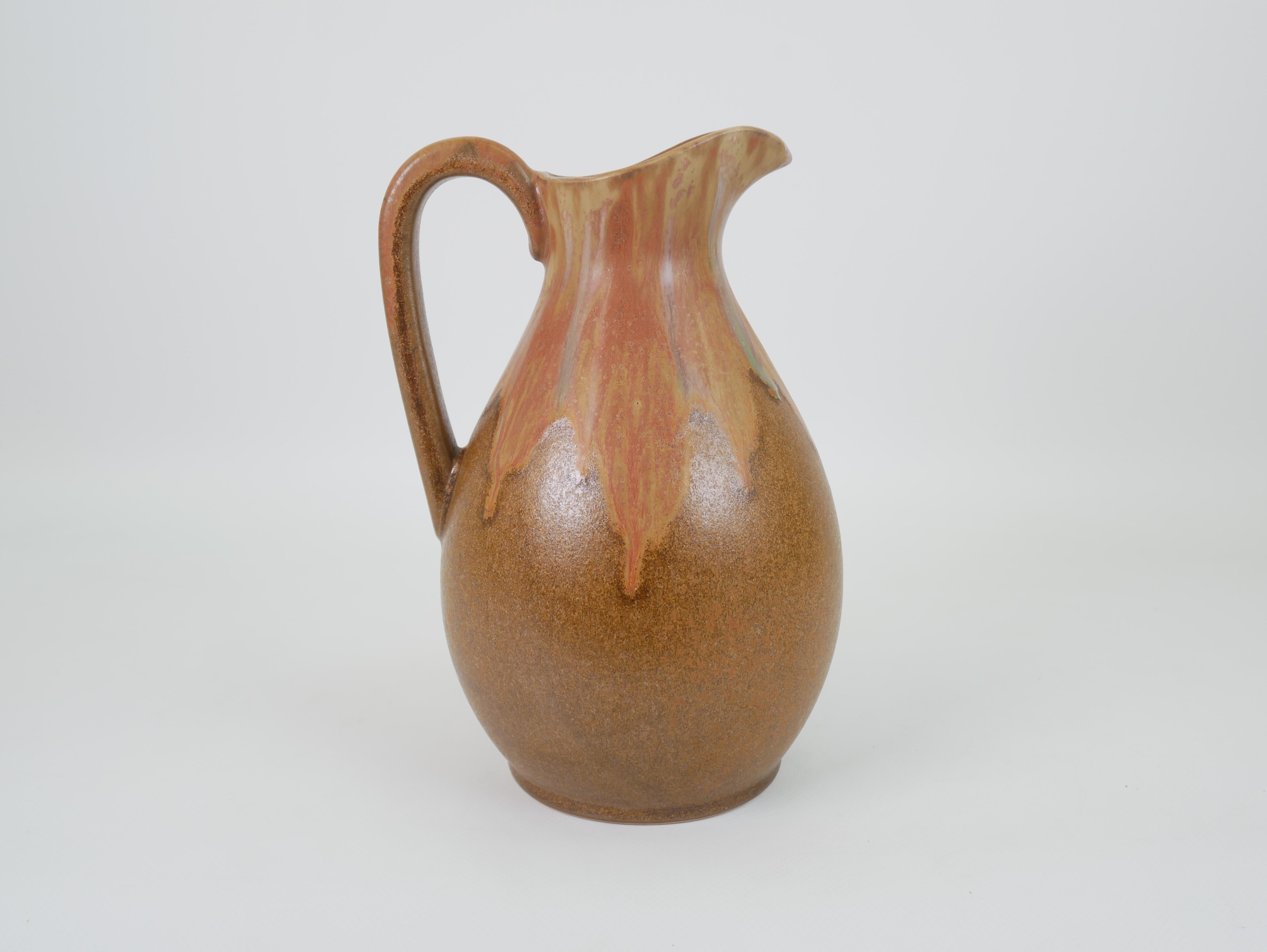 This 23 cm tall Art Nouveau pitcher is a striking piece from the Denbac ceramics manufactory, renowned for its organic forms and flambé glazes. The pitcher features a bulbous body with a high-arching handle and elegantly flared spout. Its surface is