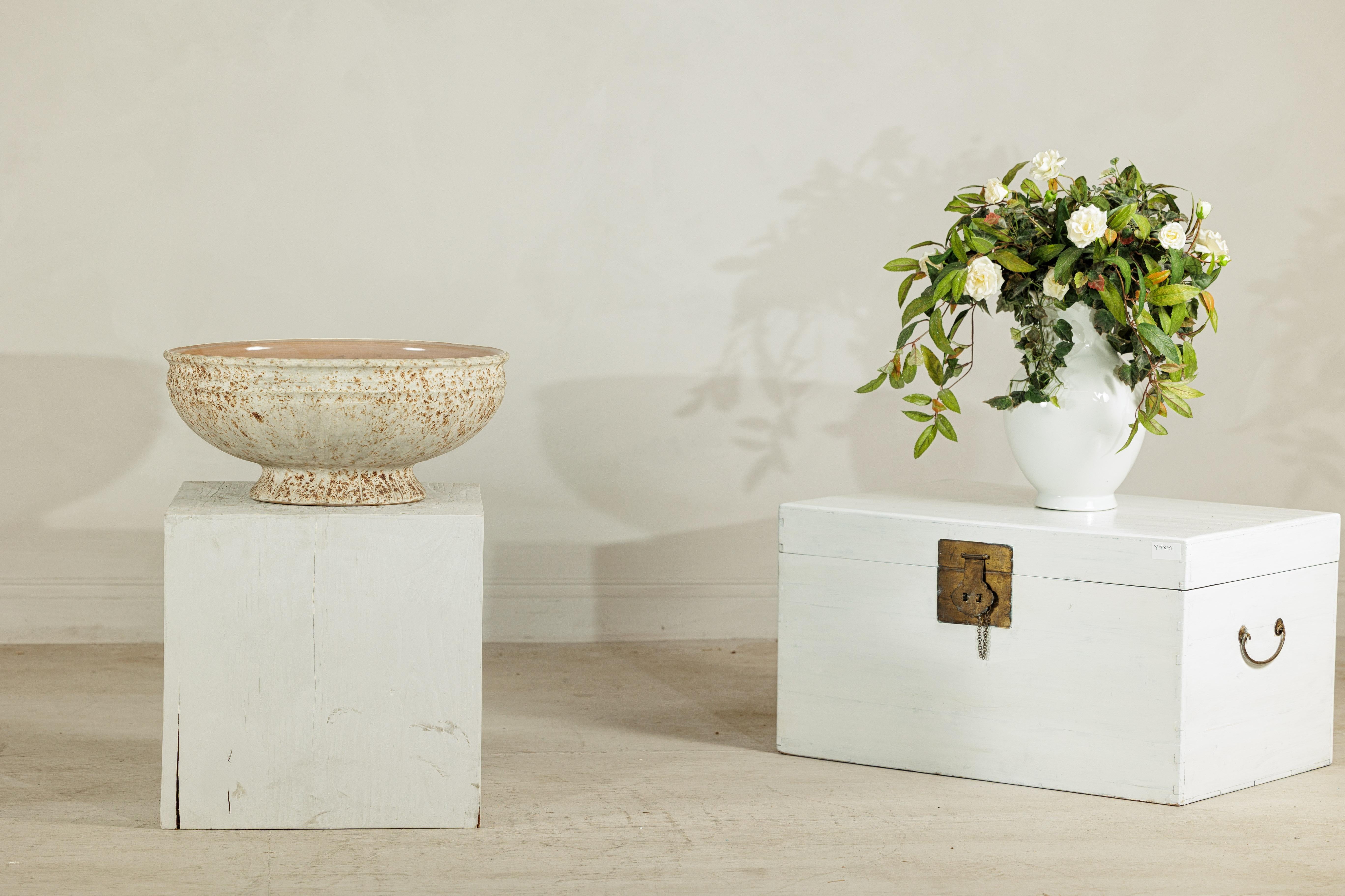 This large artisan ceramic bowl combines organic texture with refined form, creating a versatile statement piece that bridges rustic and contemporary design. The wide, open bowl rests on a raised circular base, lending the piece an architectural