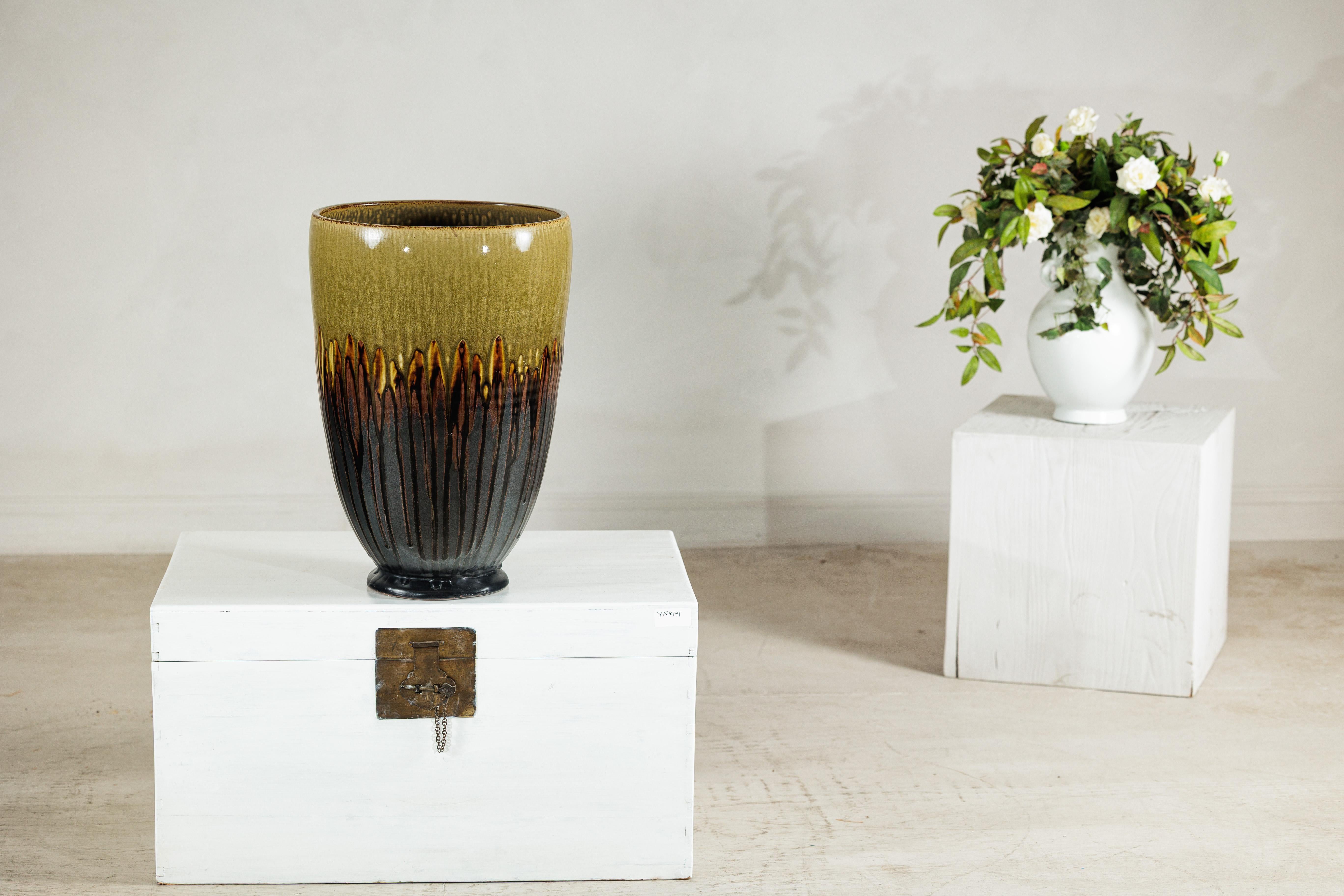 Questa grande fioriera artigianale in ceramica è caratterizzata da un sorprendente smalto stratificato che passa dal verde oliva opaco al marrone lucido e ai toni del carbone profondo. La superficie è arricchita da uno scenografico effetto a goccia