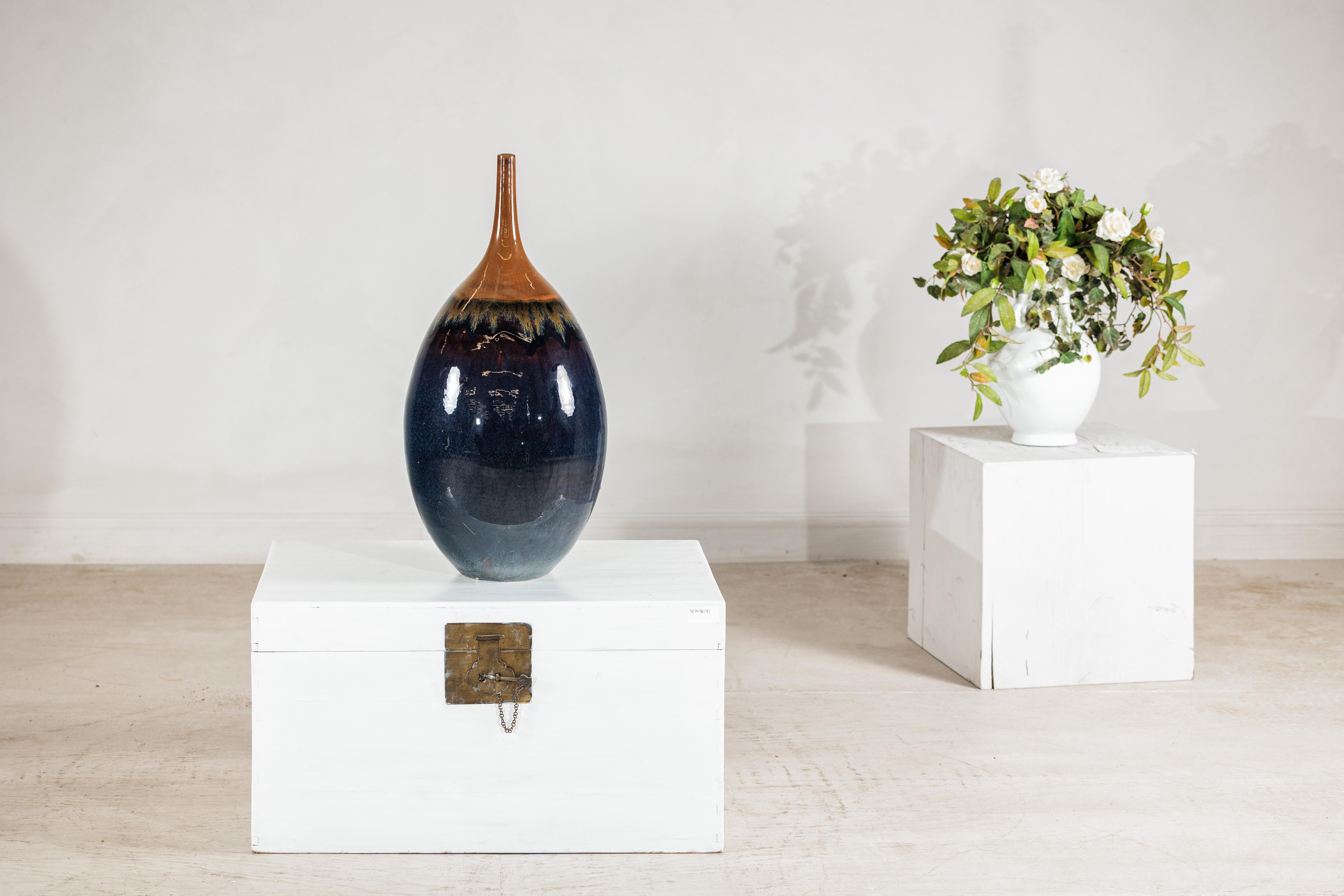 Questo grande vaso in ceramica lavorato artigianalmente presenta un sorprendente studio di proporzioni e smalti. Il collo allungato e affusolato, in una calda tonalità marrone, si trasforma in un corpo ovoidale rifinito in un profondo blu notte. I