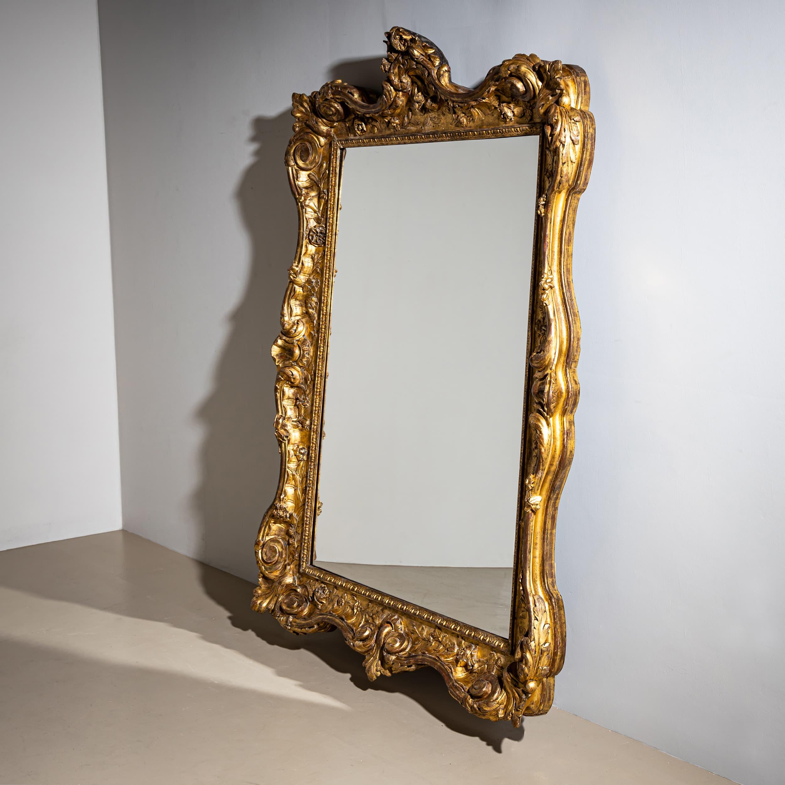 Impressive Baroque wall mirror dating to the mid-18th century, featuring an elaborately gilded frame with double volutes in the corners, finely carved rose garlands, and a decorative floral crest at the top. The mirror glass has been replaced. 