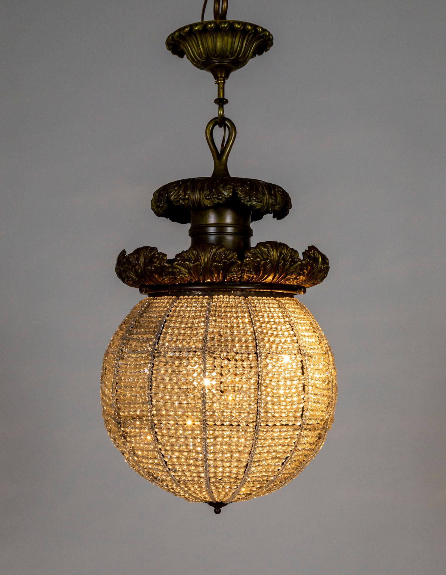 A sizable, crystal ball pendant light, topped with giant, cast bronze, curling acanthus leaves. It is composed of innumerable strands of faceted crystal beads wired onto a large, orb shaped armature that acts as a shade to a 3-socket cluster. The