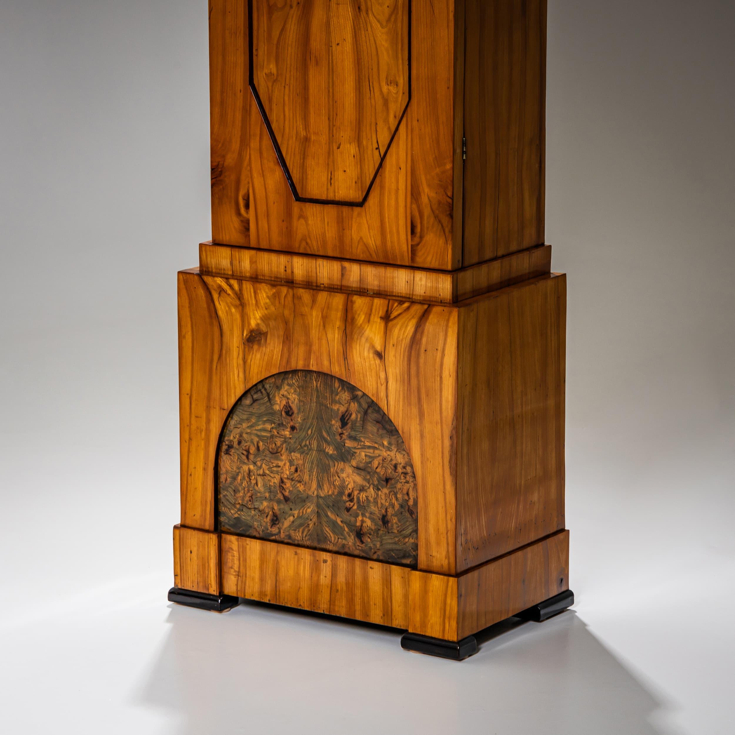 Elegante orologio a cassa lunga Biedermeier con cassa in ciliegio finemente impiallacciata. La sottile sezione centrale presenta una porta con un pannello ottagonale. La base è evidenziata da un segmento verde-patinato, a cui fa eco una stretta