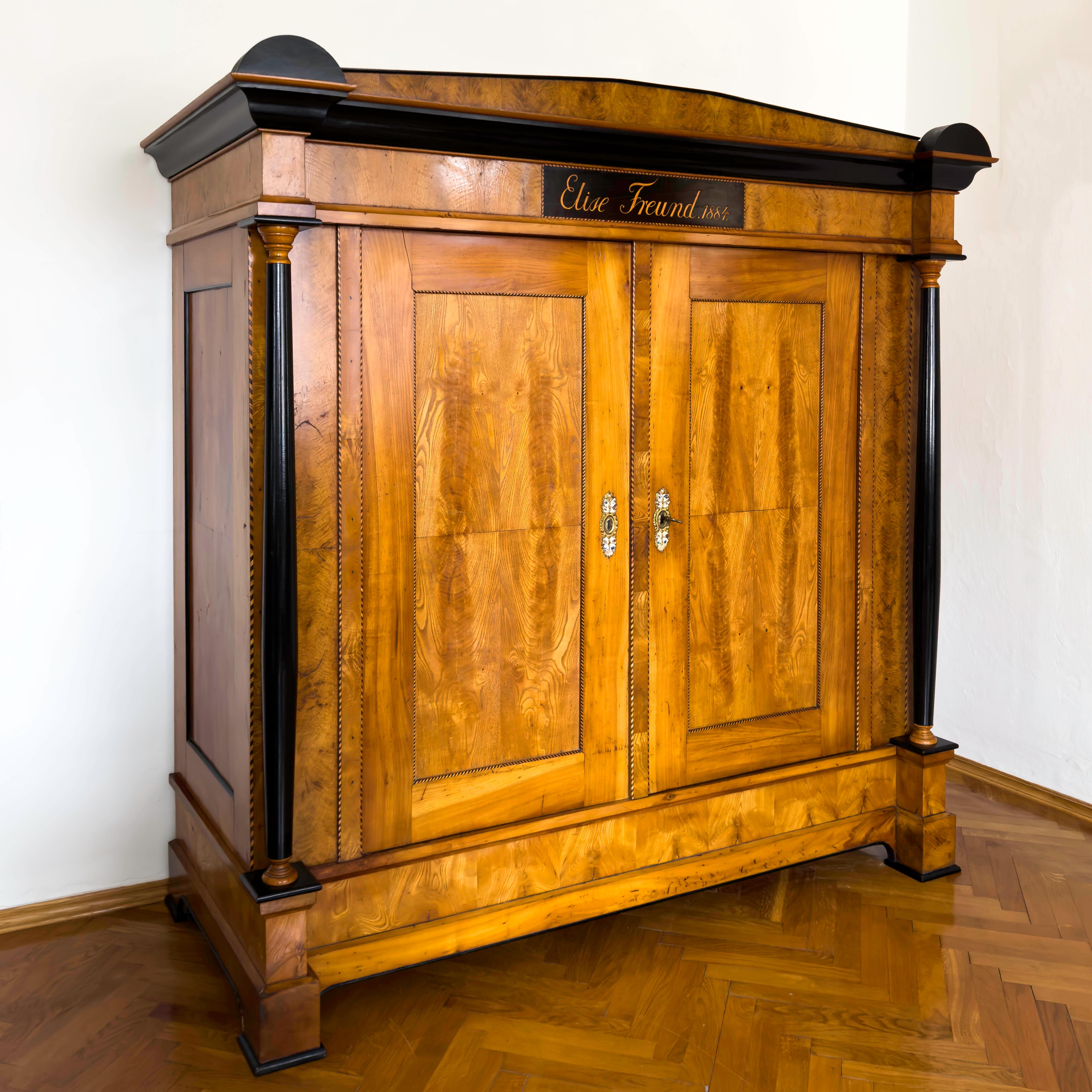Large two-door wardrobe in ash and cherry with fillet band inlays and ebonised columns. The cornice with accentuated pediment and ebonised segmental elements is inscribed: Elise Freund, 1884. The sides are coffered and framed with ebonised profiles.