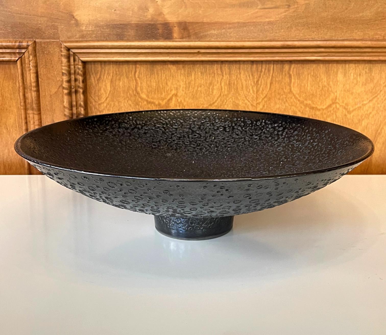 Un bol sur pied en céramique de taille impressionnante, réalisé par le potier californien James Lovera (1920-2015), probablement au début des années 2000. Le bol est entièrement recouvert d'une glaçure de lave métallique noire, qui a fait la
