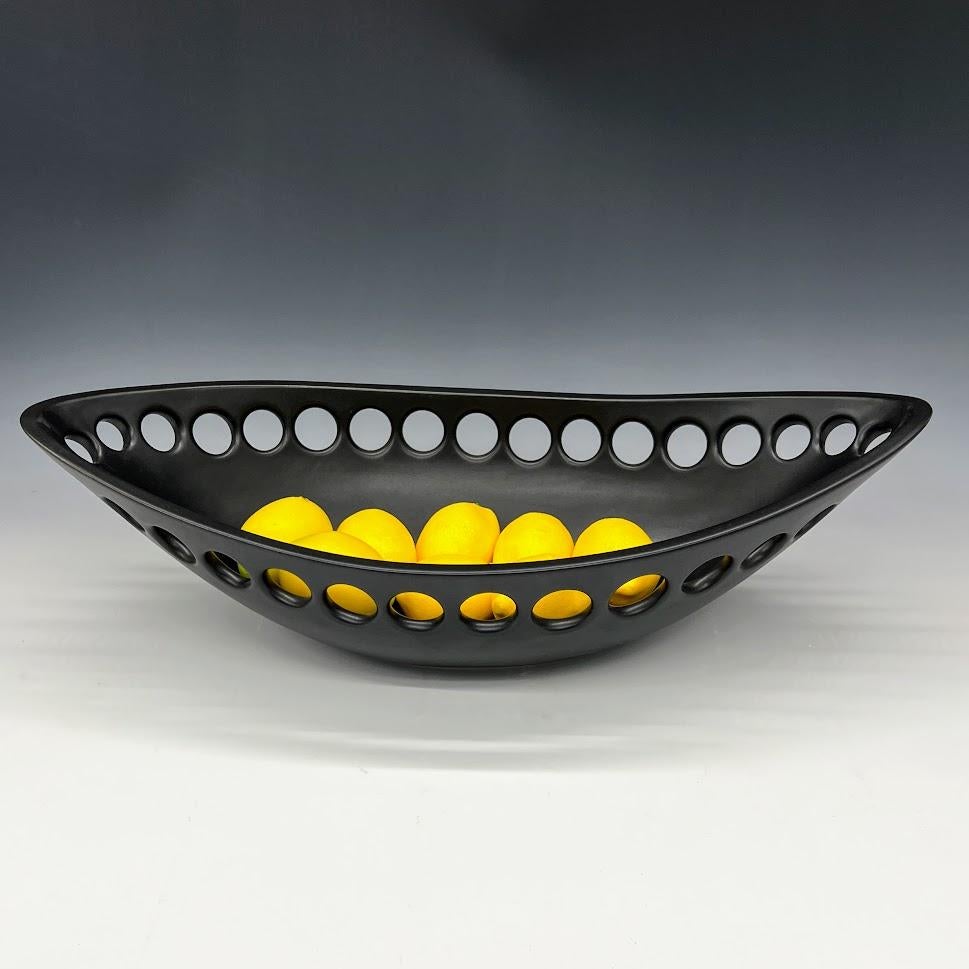Inspired by organic contemporary design, this pierced oblong bowl is wheel thrown, distorted and hand pierced stoneware with a black satin glaze. Small holes are created when the clay is still wet and then each hole is painstakingly enlarged and