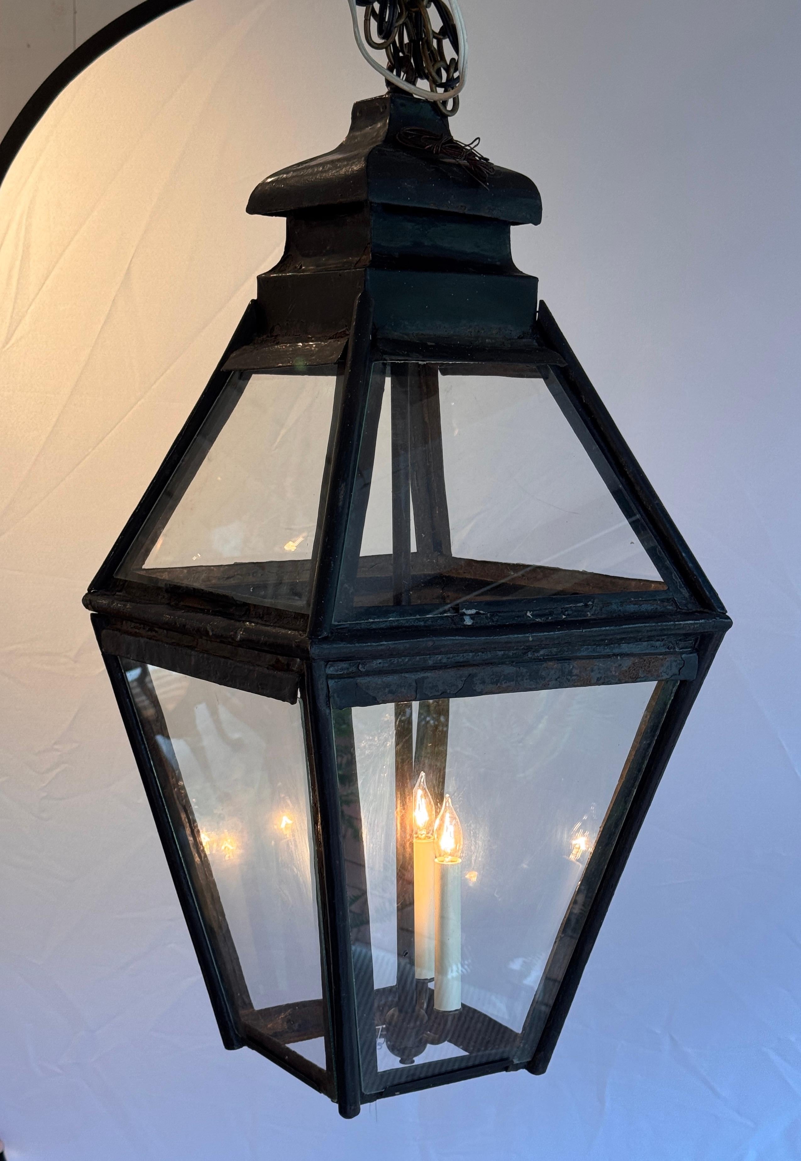 Substantial black-painted lantern with classic tapered sides and clear glass panels. Features a double candle-style light fitting and open base construction. The patina and scale suggest traditional craftsmanship, ideal for covered entries or