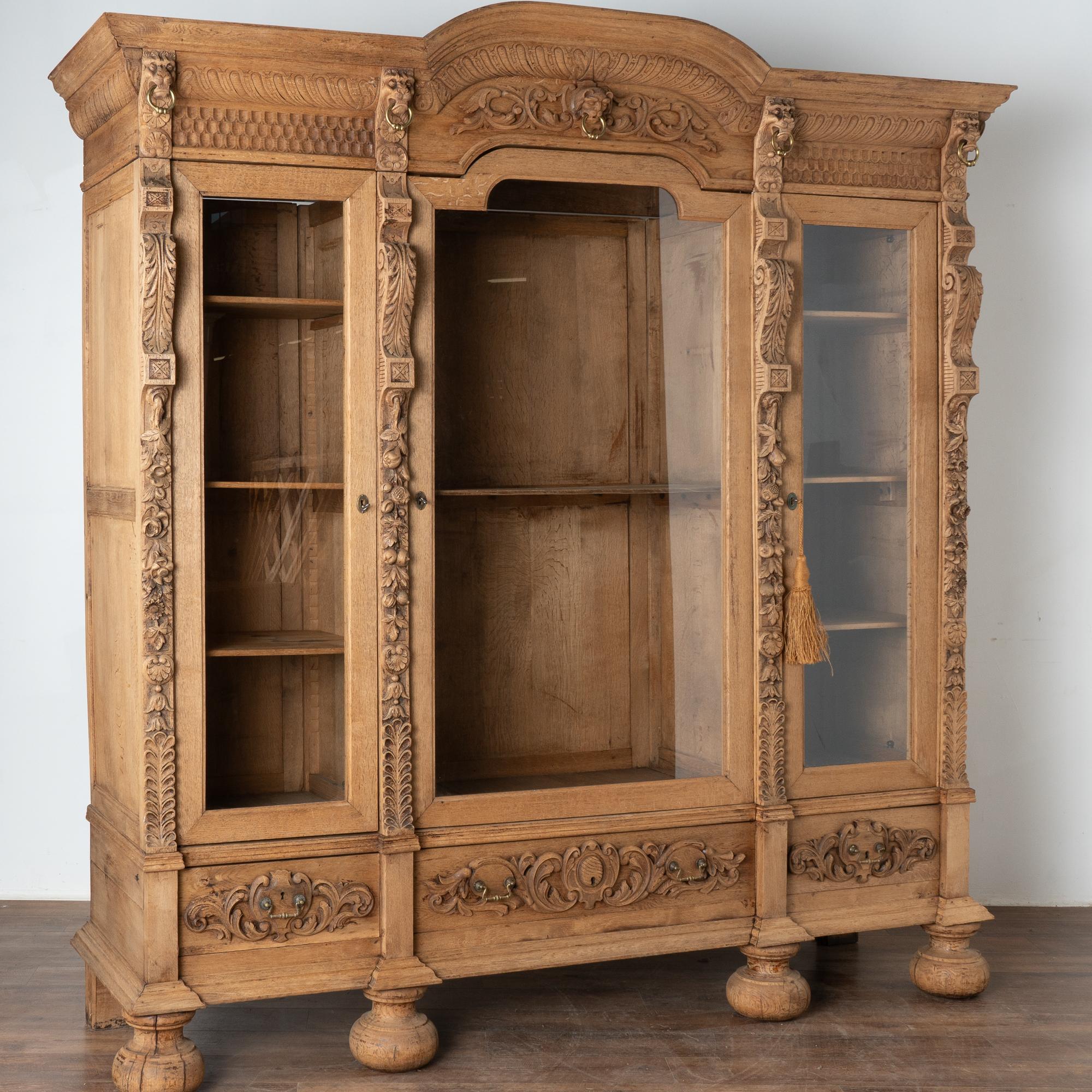 This dramatic bleached oak bookcase or display cabinet has three long glass panels and stands 8' tall.
Notice the highly carved columns, crown (with lion heads), and 3 drawer fronts along the lower section. 
Ideal for display of books or any