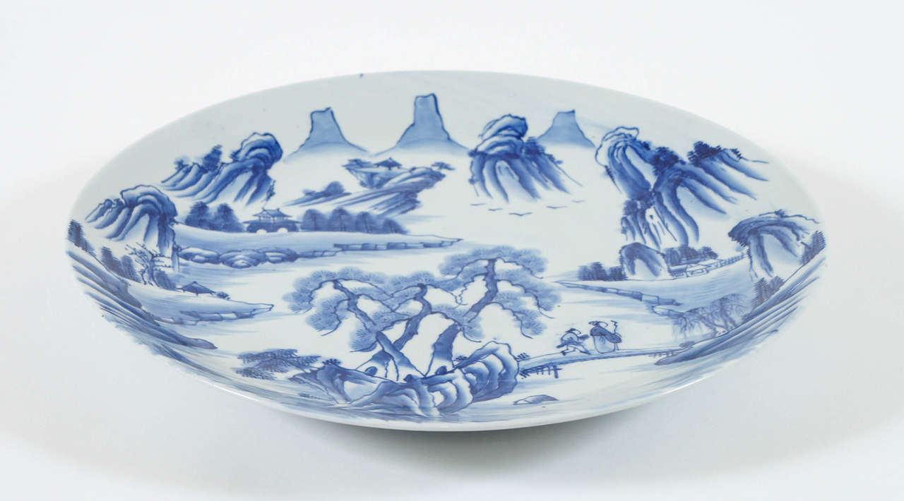 Chargeur de grande taille en porcelaine bleue et blanche de l'époque Meiji (Japon), datant d'environ 1880. Il présente un paysage de montagne peint sous glaçure en chinoiserie avec deux personnages traversant un pont.