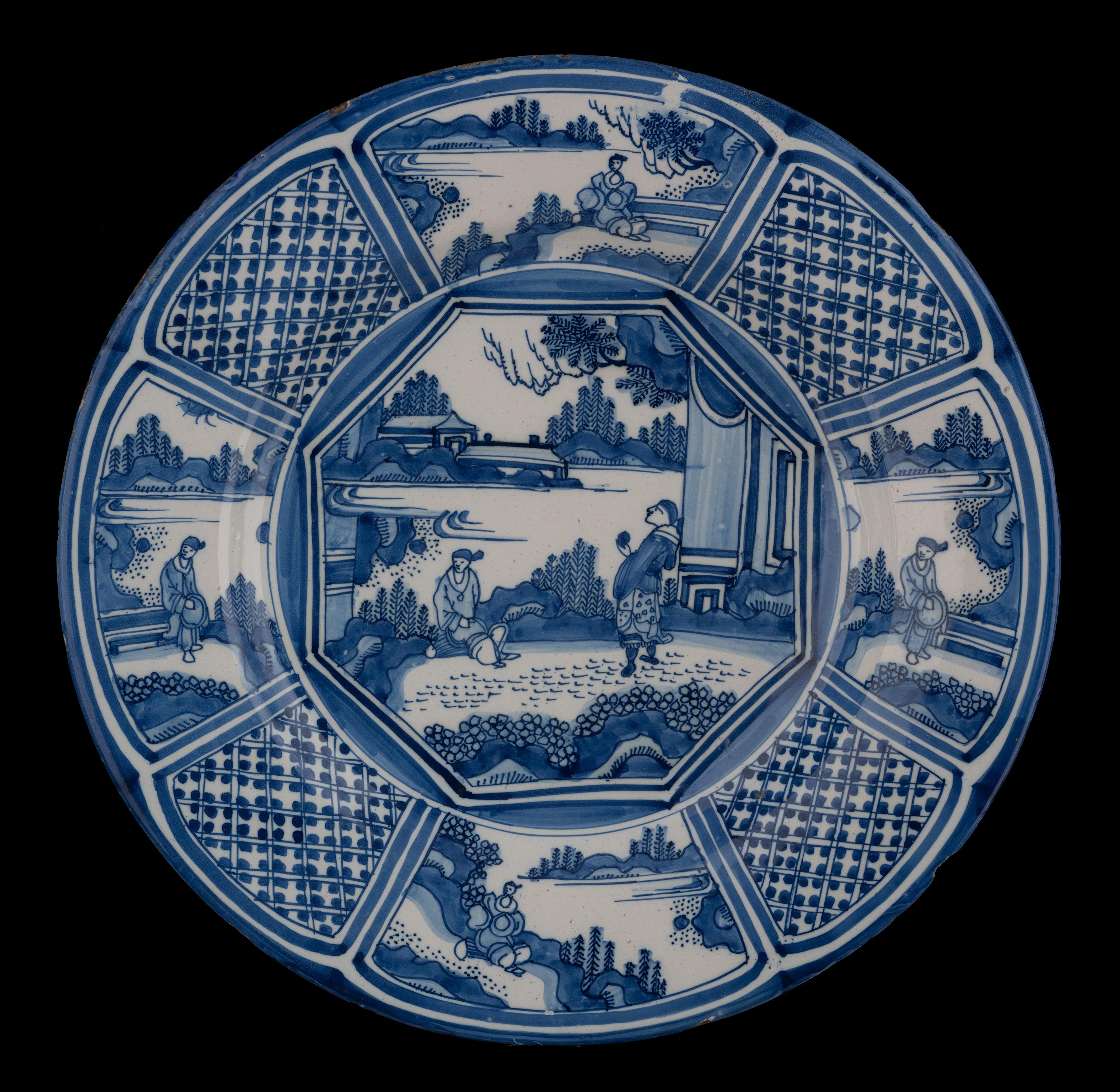 The blue and white dish has a wide, flat flange and is painted in the centre with two Chinese figures on a terrace in an oriental garden landscape. Two pavilions are visible in the background. The scene is framed in an octagon. The well and border