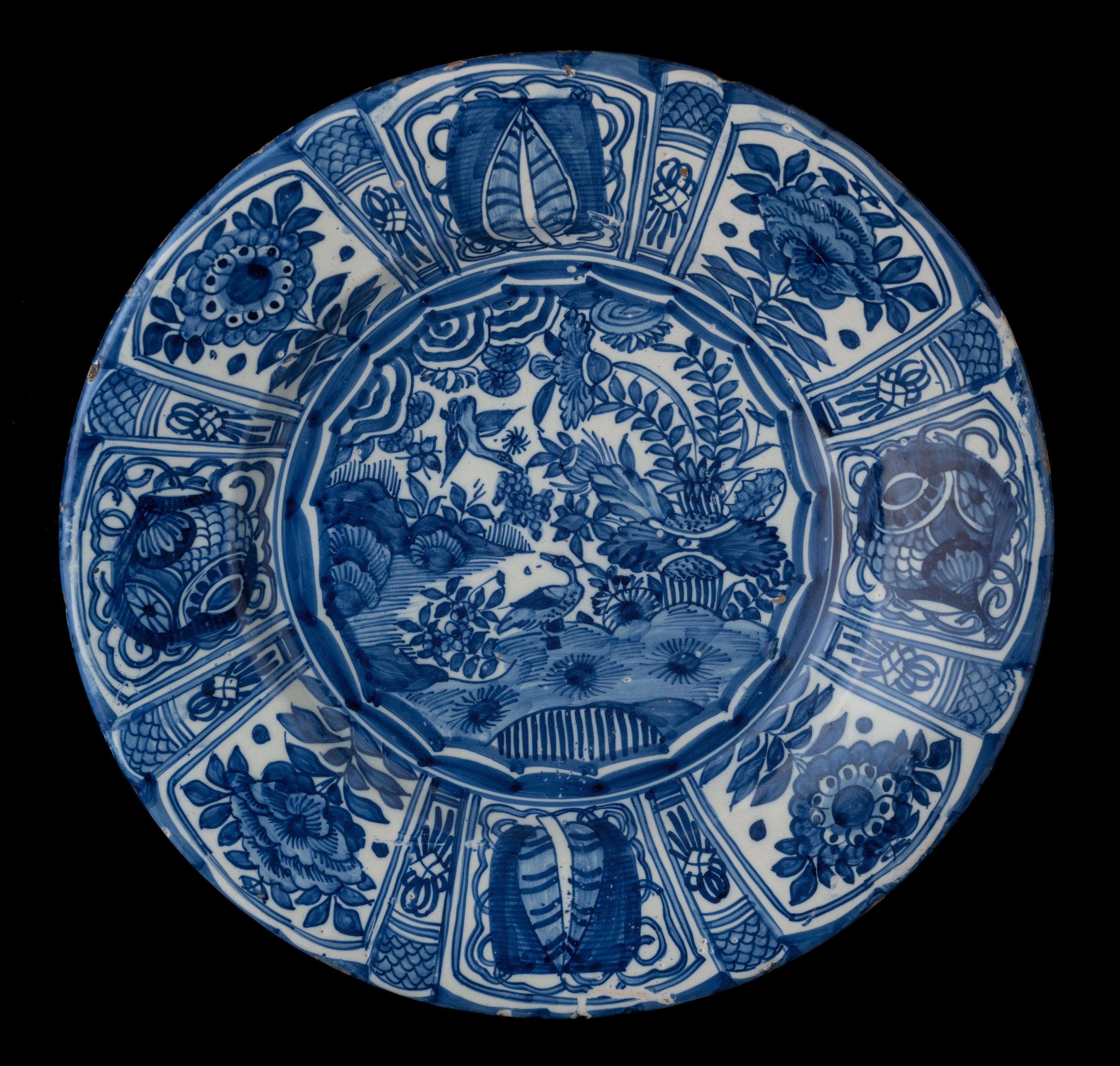 The blue and white dish has a wide, spreading flange and is painted in the centre with a Chinese-style rocky landscape with plants, flowers and birds. The depiction is framed within a polygon shape. The well and flange are divided into eight wide
