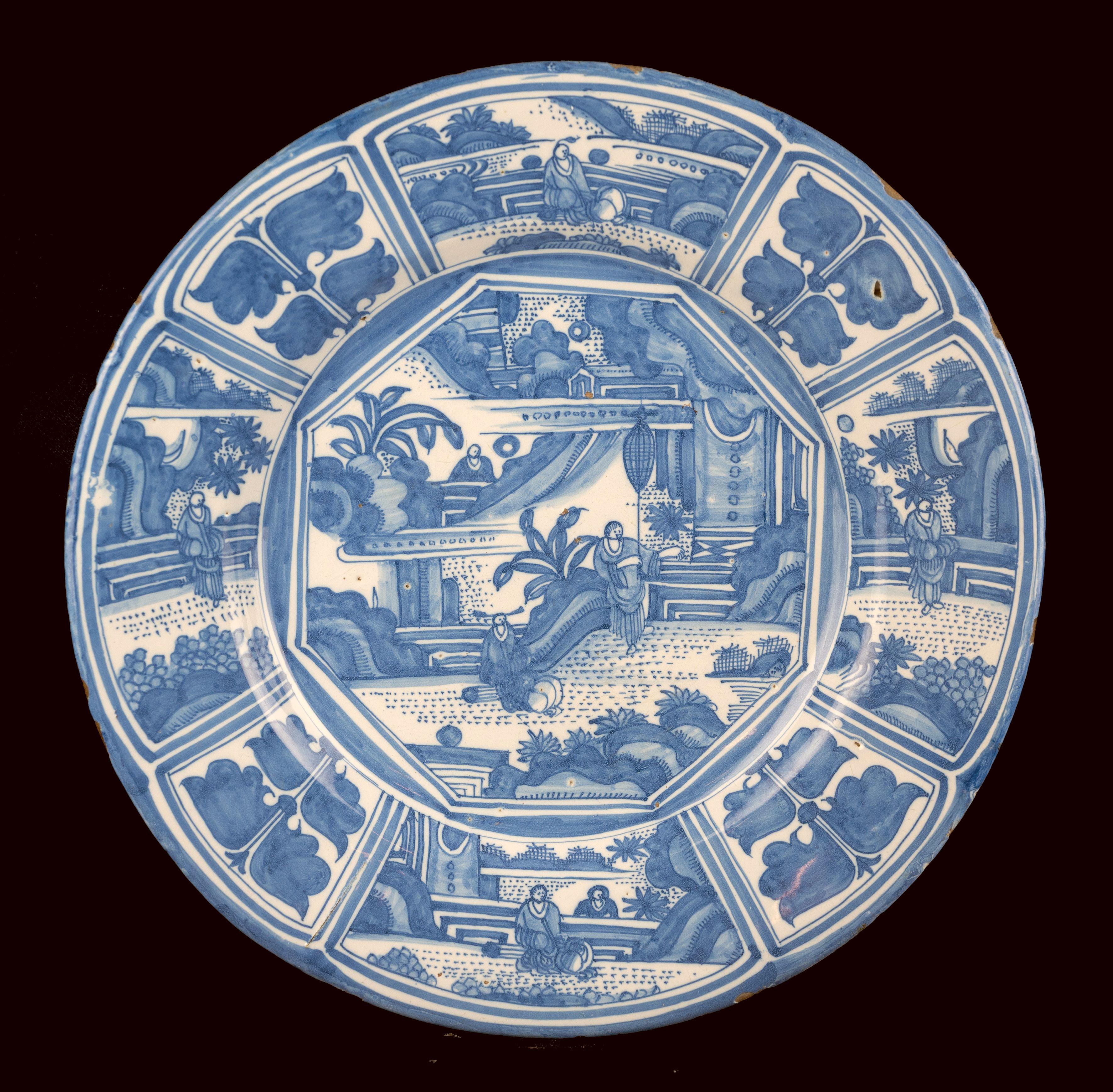 The blue and white dish has a wide-spreading flange and is painted in the centre with two Chinese figures on a terrace by a pavilion in an oriental garden landscape. A third Chinese is visible in the background. The scene is framed in an octagon.