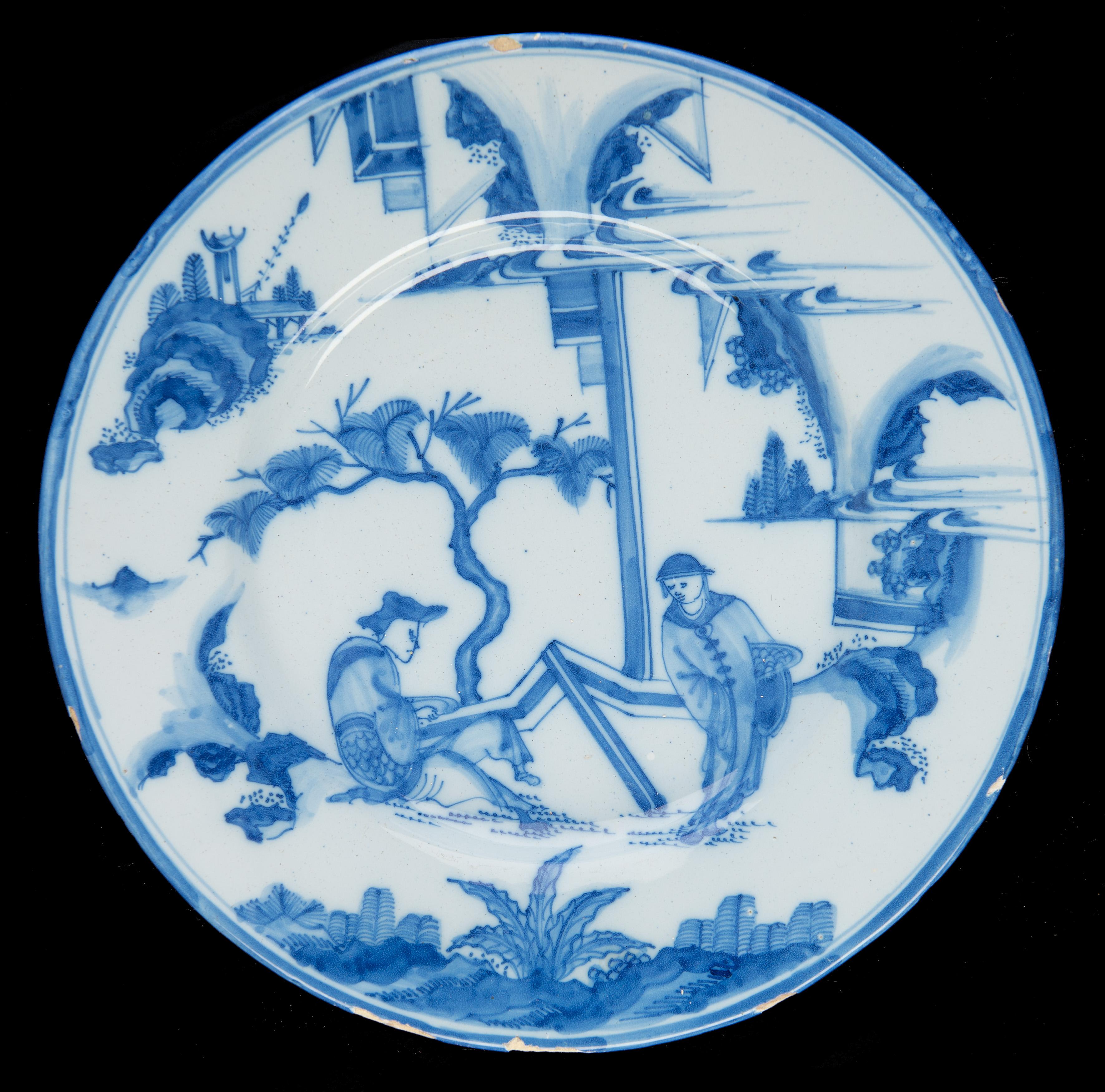 Dish with a wide-spreading flange, the surface painted entirely with a blue and white chinoiserie decoration of two men in a garden-like landscape. Both men carry a dish, and on the dish of the standing man lies round fruit. The rim is executed in