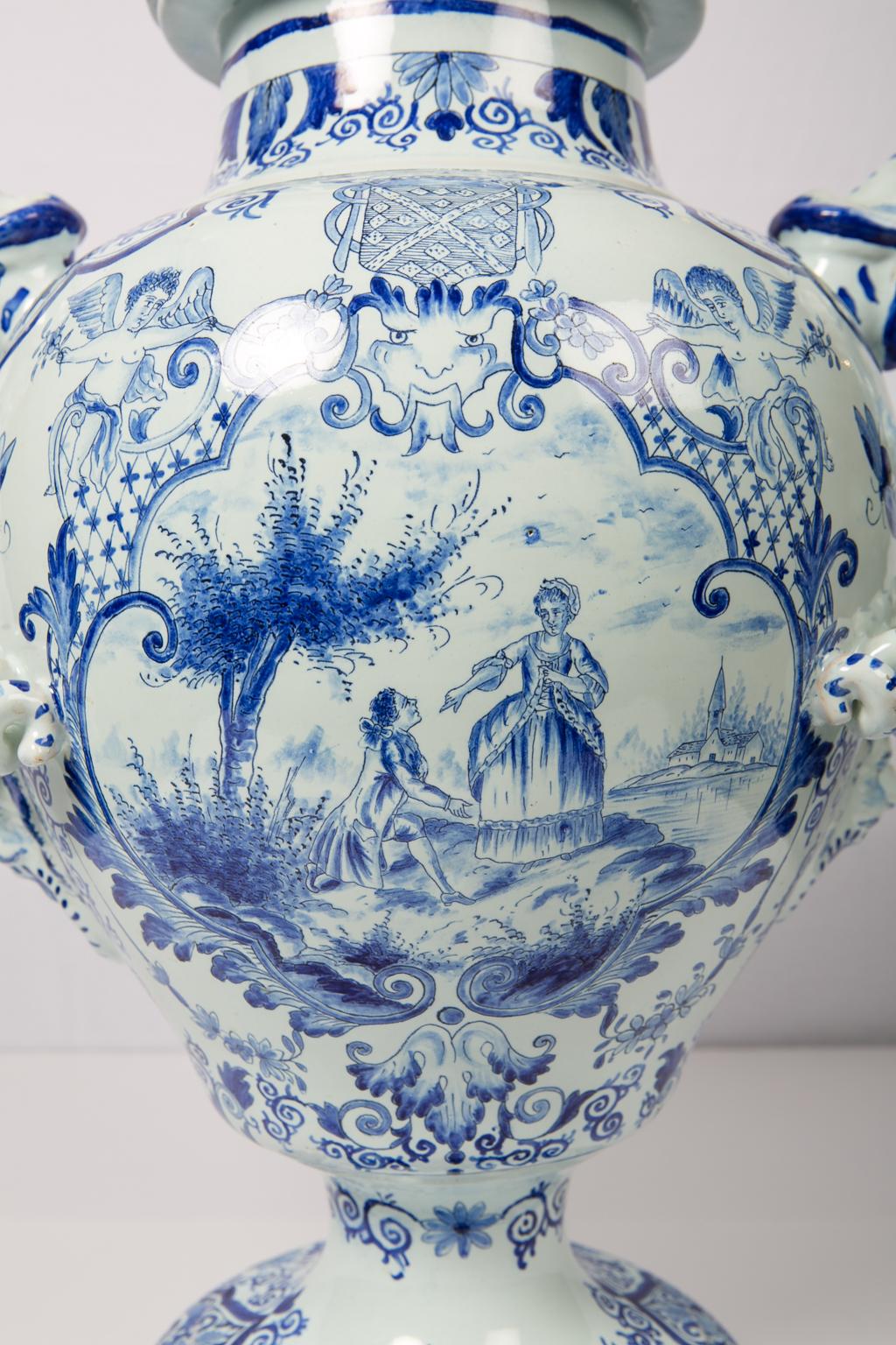 A large Dutch delft blue and white covered urn decorated in several shades of blue featuring an armorial shield supported by angels. The striking handles are in the form of coiled snakes. The base, shoulder, and cover of the vase are decorated with