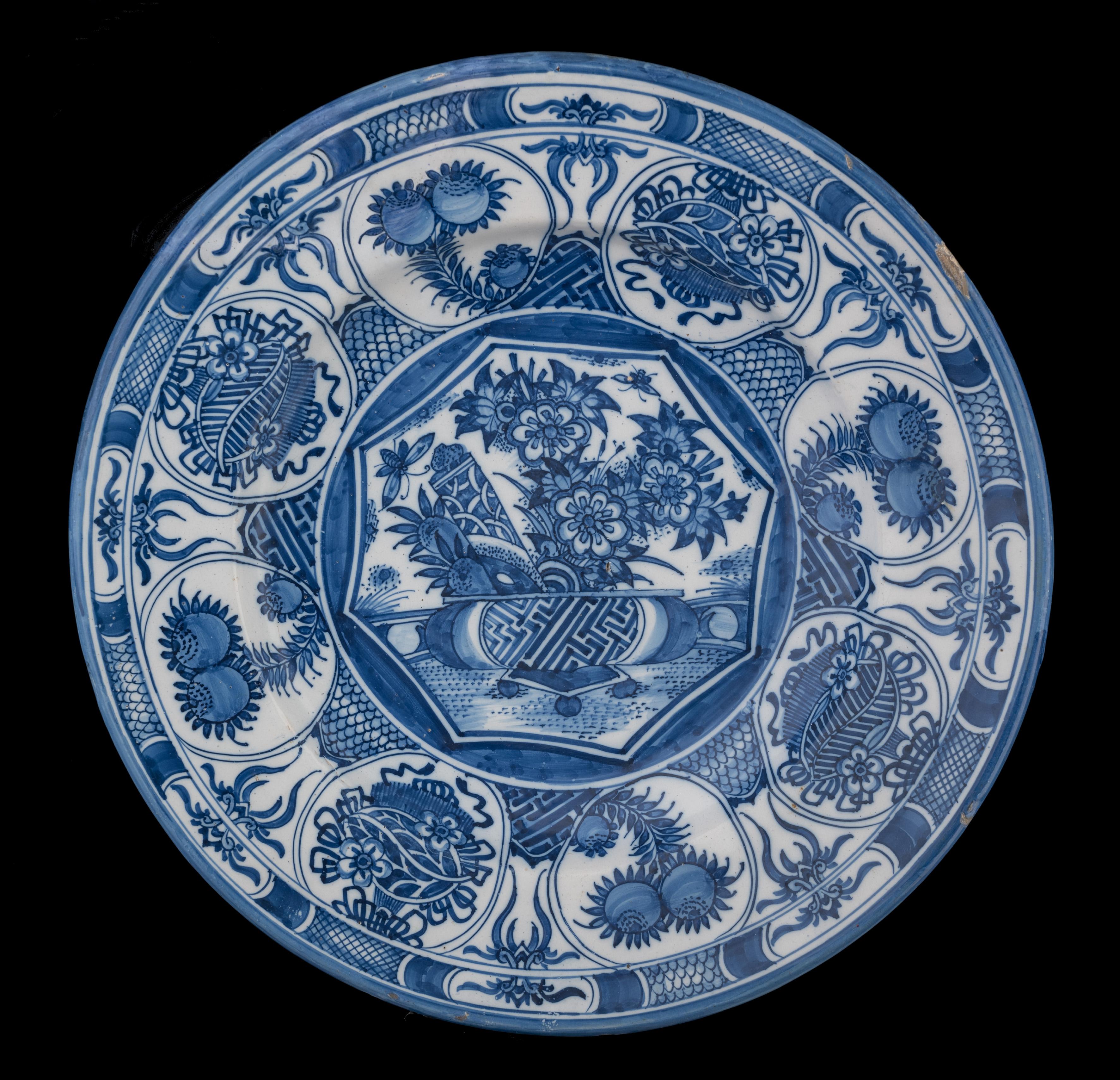 The blue and white dish has a wide-spreading flange. The centre is painted with a basket on a low square table, filled with flowers, fruits, a book scroll and insects. It is placed on the ground in front of a balustrade. The basket is decorated with