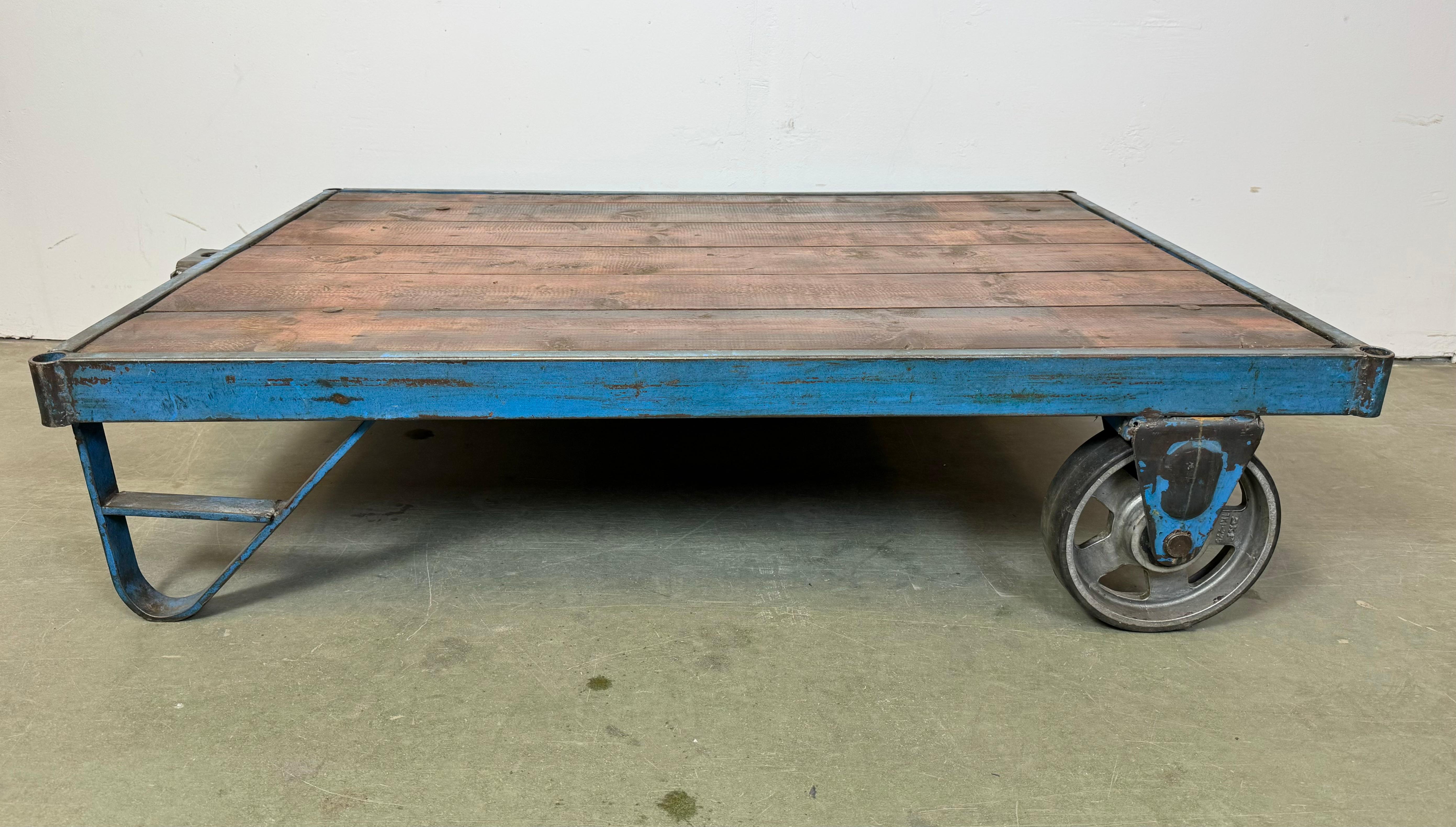 Ein ehemaliger Gabelhubwagen aus einer Fabrik dient jetzt als Couchtisch. Er hat eine blaue Eisenkonstruktion mit zwei Originalrädern und einer massiven Holzplatte. Das Gewicht des Tisches beträgt 40 kg.