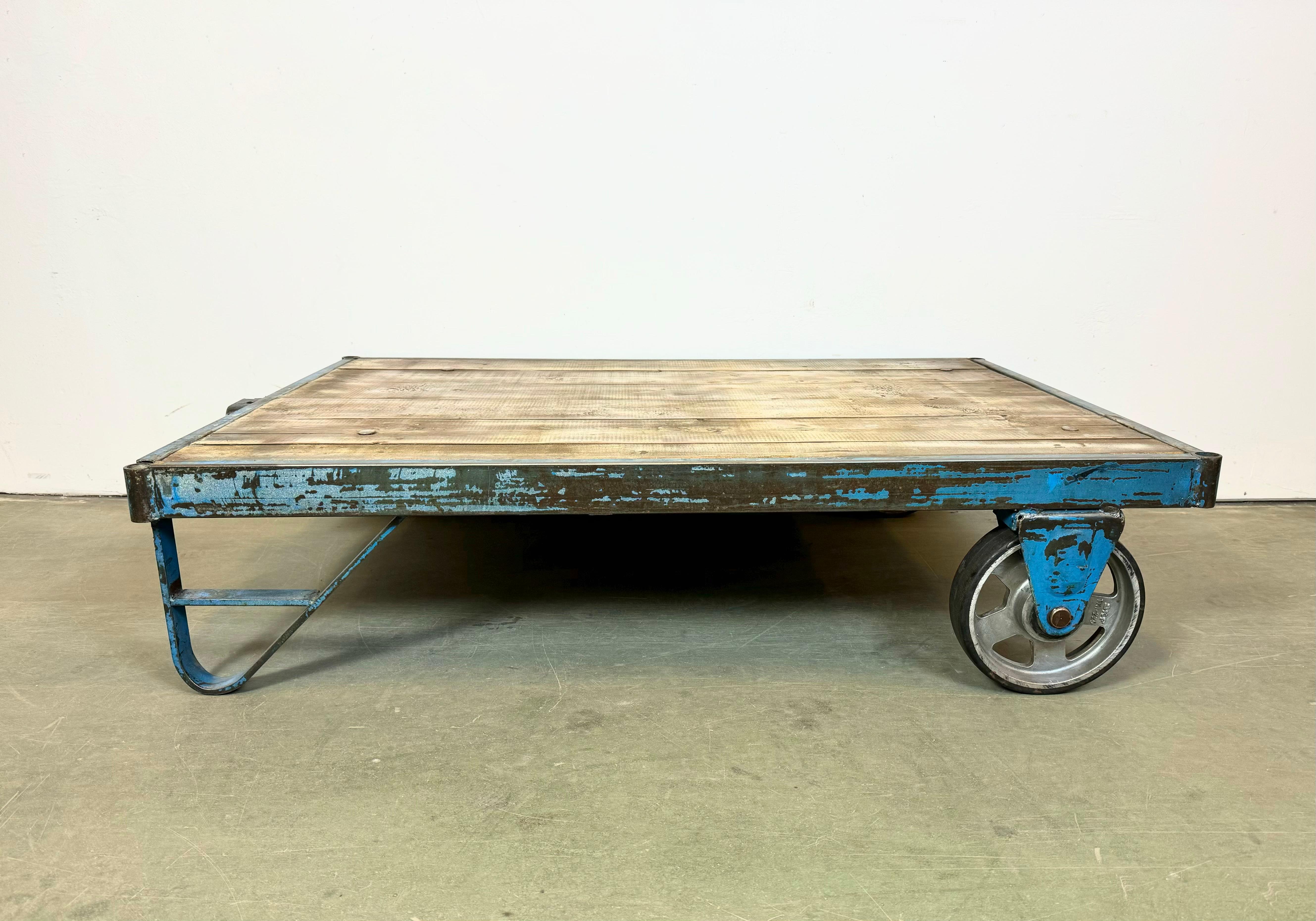 Former pallet truck from a factory now serves as a coffee table. It features a blue iron construction with two original wheels and solid wooden plate. The weight of the table is 40 kg.