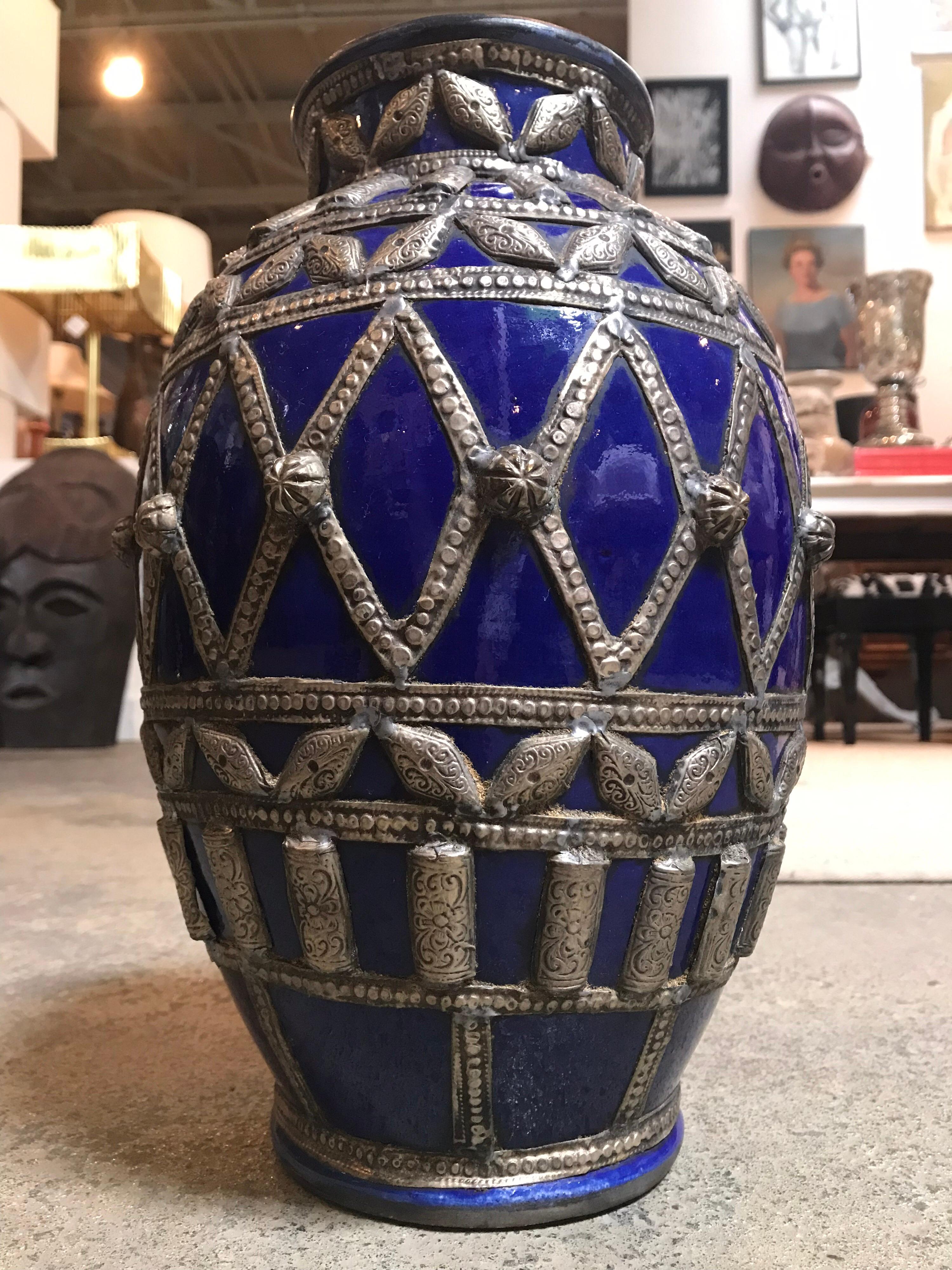 This larger, dark blue Moroccan pot is hand painted and has a ceramic glaze. It is decorated with various ribbons of silver-like metal providing round geometric patterns throughout.