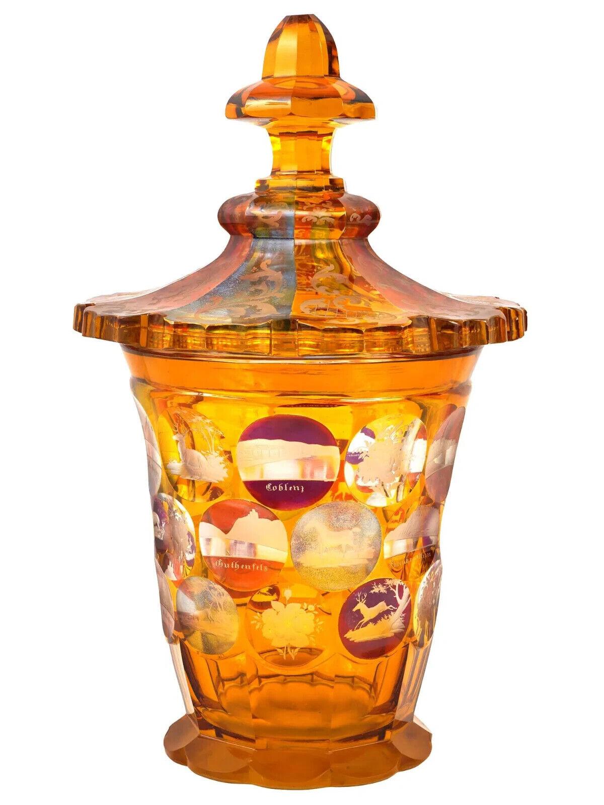 Un grande vaso in vetro di Boemia color ambra con coperchio abbinato. Il vaso ha un corpo alto e leggermente svasato, decorato con pannelli ovali in vetro smerigliato inciso che raffigurano paesaggi, edifici e animali. Il coperchio è a cupola e
