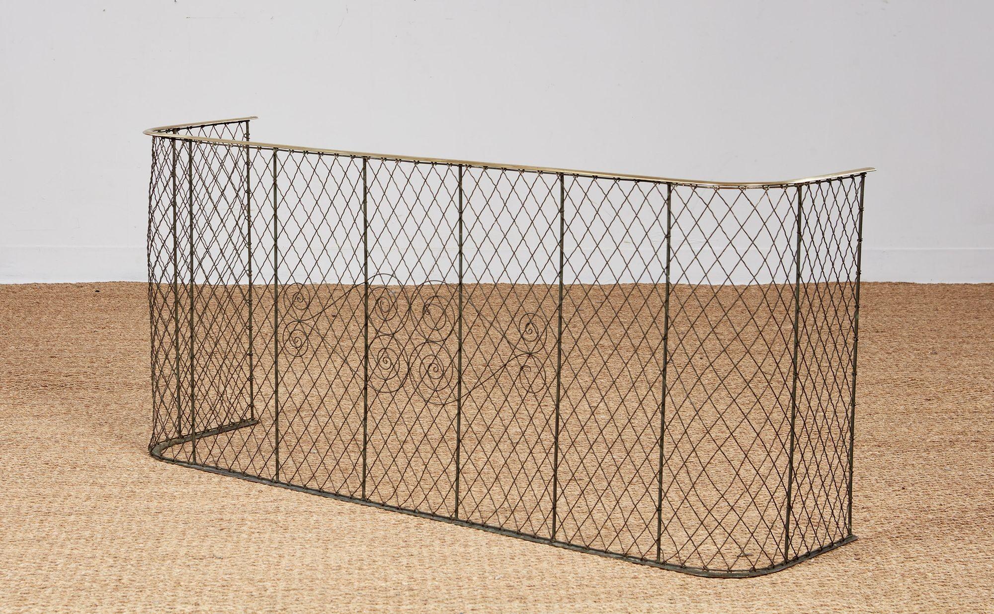 A fireplace screen with diagonal wide mesh on ten vertical iron supports and with an iron base rail and a brass top rail, with iron scrollwork to the center. This one is particularly long (53
