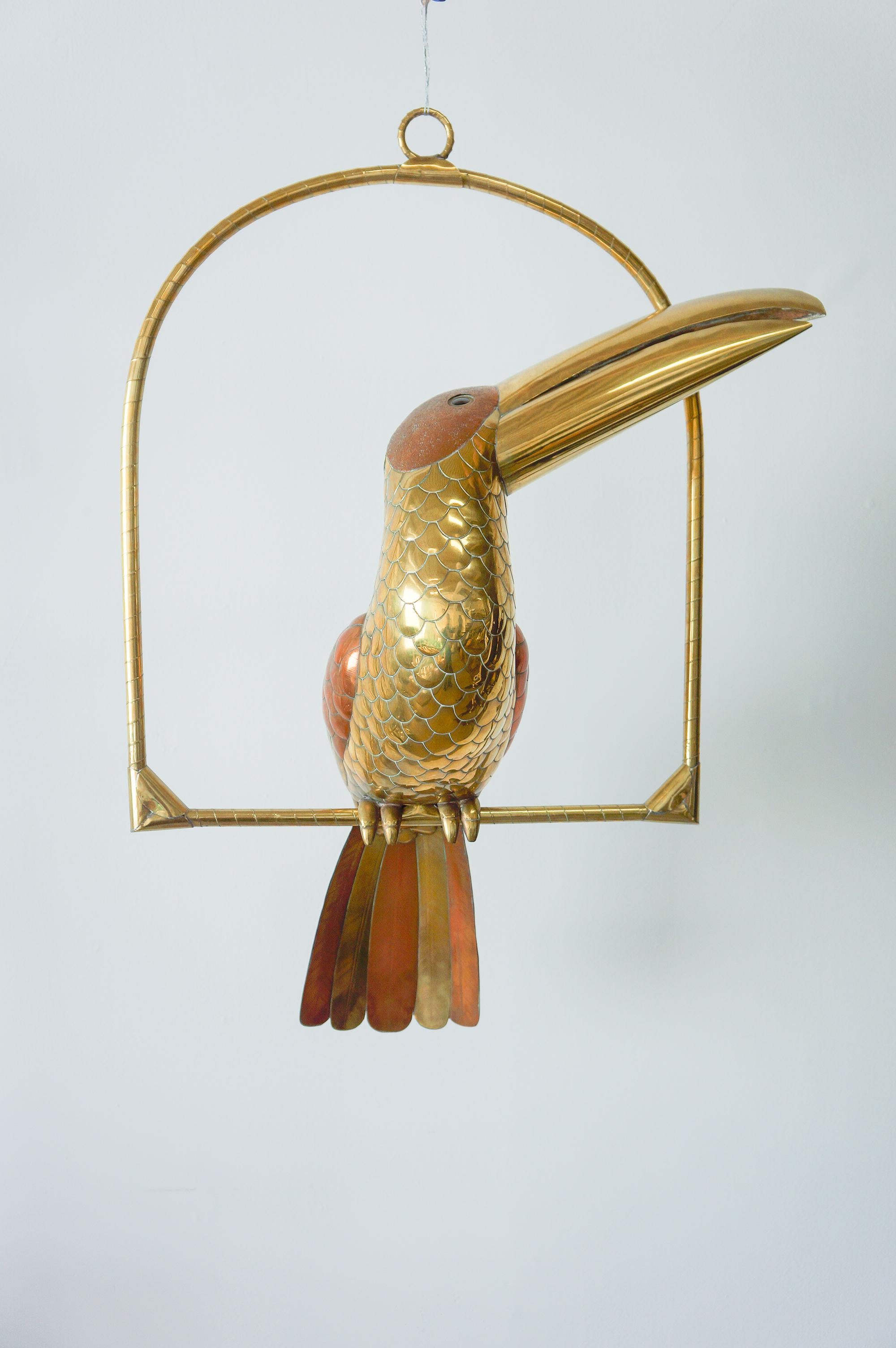 Large, life-sized brass and copper toucan on a hanging perch handmade by Sergio Bustamante. 

The world-renowned Mexican artist started creating unique animal themed metal sculptures in the 1970s. Sergio Bustamante, a luminary in the realm of