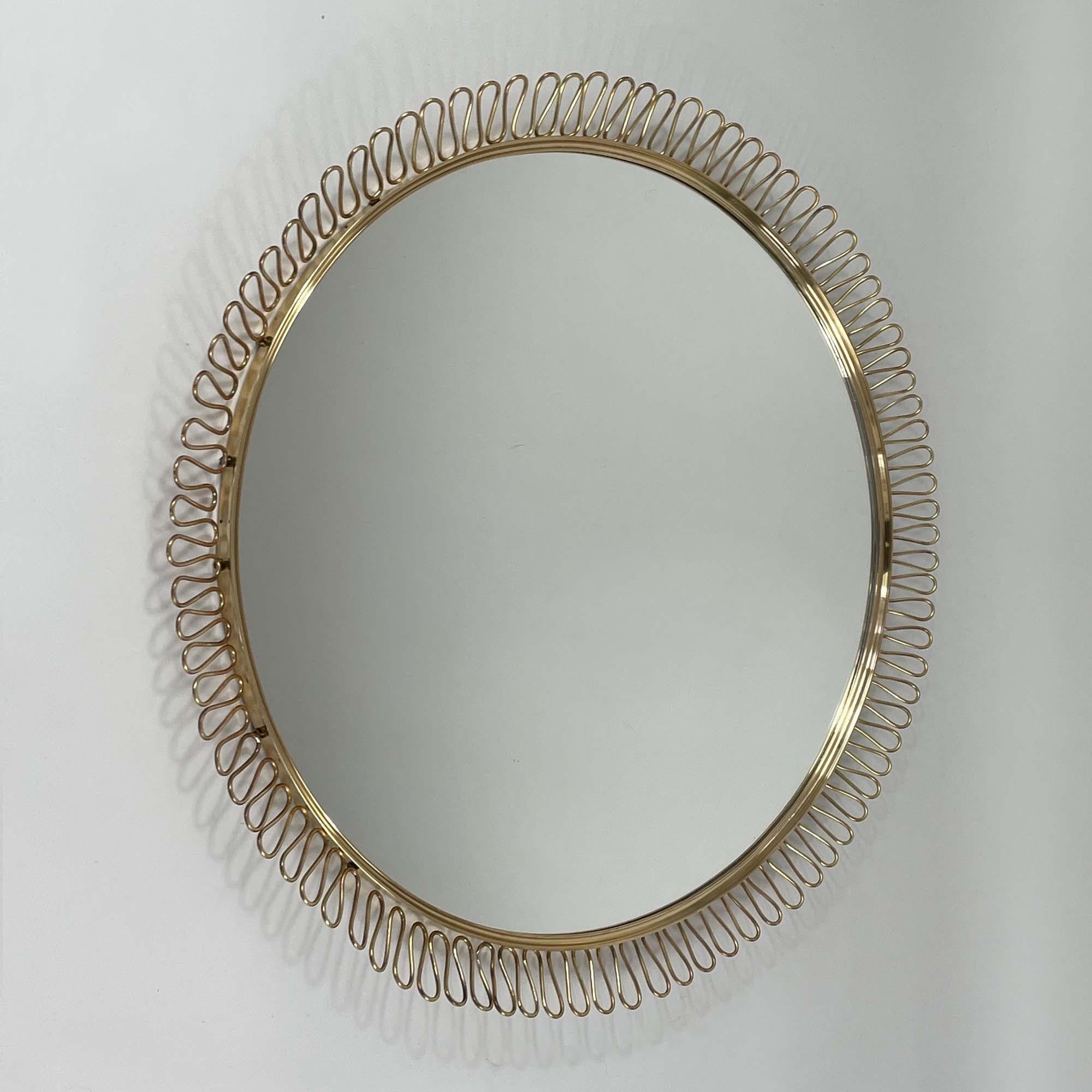 Ce miroir mural vintage de style Josef Franks a été conçu et fabriqué en Suède dans les années 1950. 

Le cadre est en laiton et présente un motif décoratif composé de boucles.  rayonnant vers l'extérieur à partir du miroir lui-même. Ces boucles
