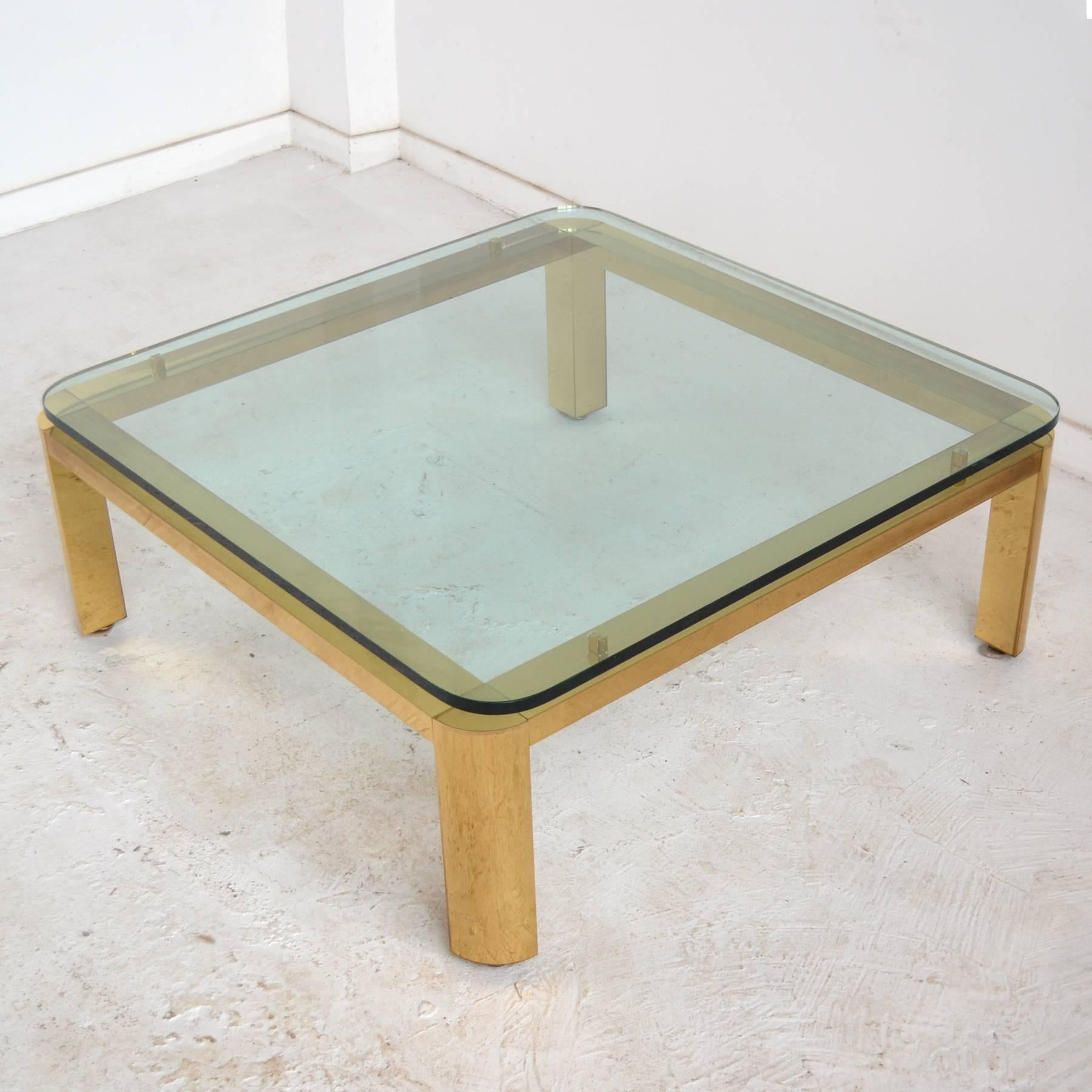 A largely scaled coffee table with an elegant brass base supporting a thick glass top with radiused corners.