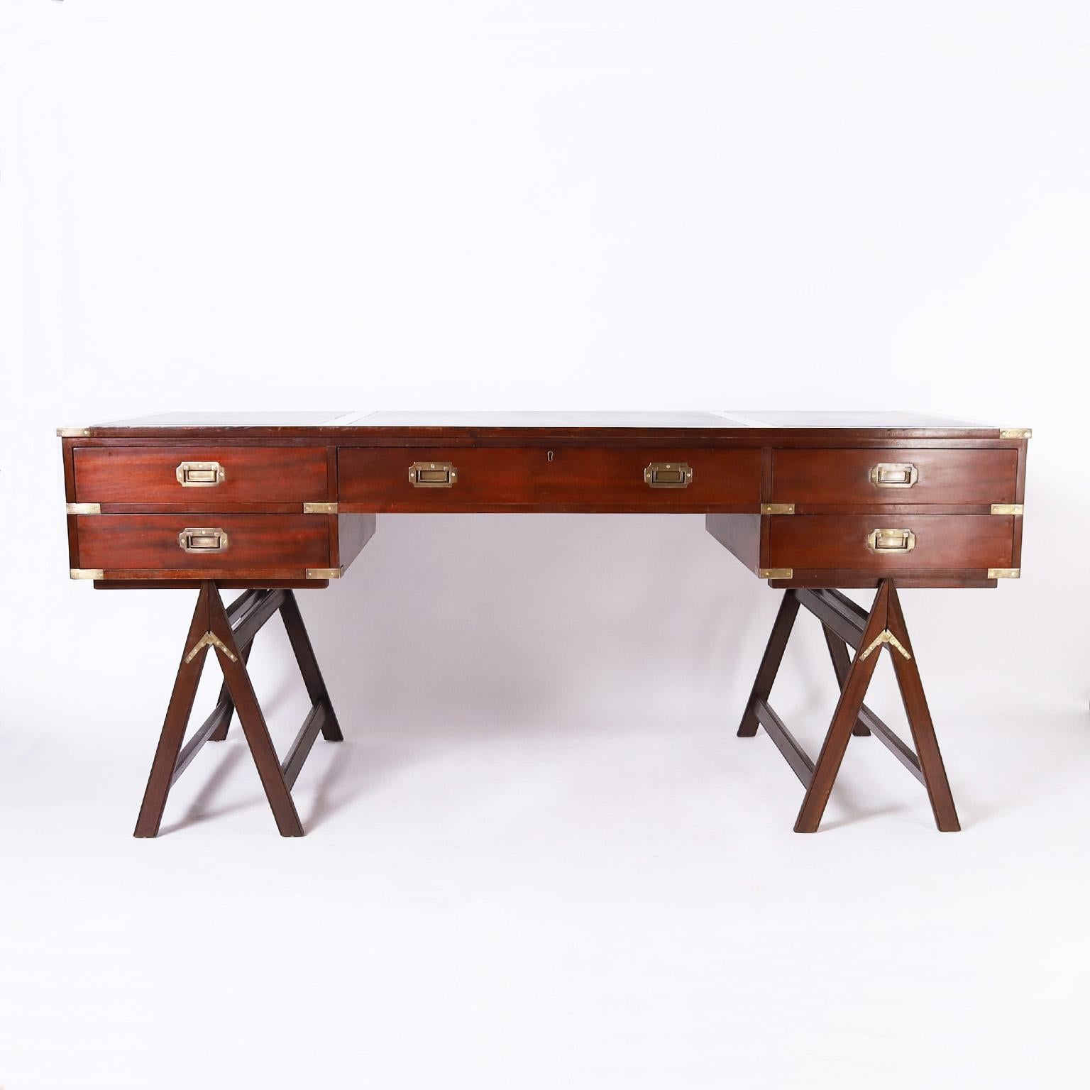 Handsome mid-century campaign style desk crafted in mahogany with a three panel black tooled leather top on a case with five drawers with brass campaign hardware on folding sawhorse style legs.