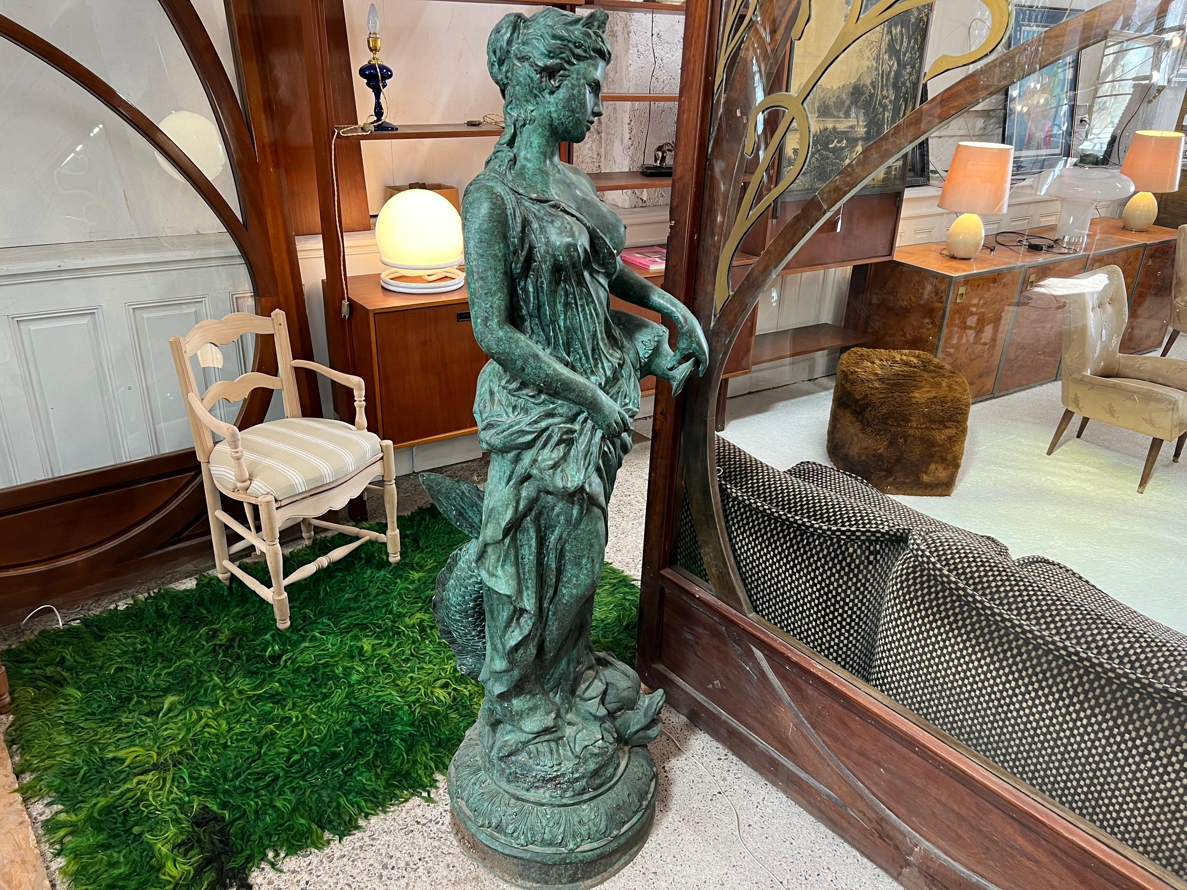 magnificent bronze statue in green bronze representing a naked woman holding an amphora in her left hand, her right hand holds her light dress
it is entwined with an antique fish at the foot on its base decorated with acanthus leaves
the statue is
