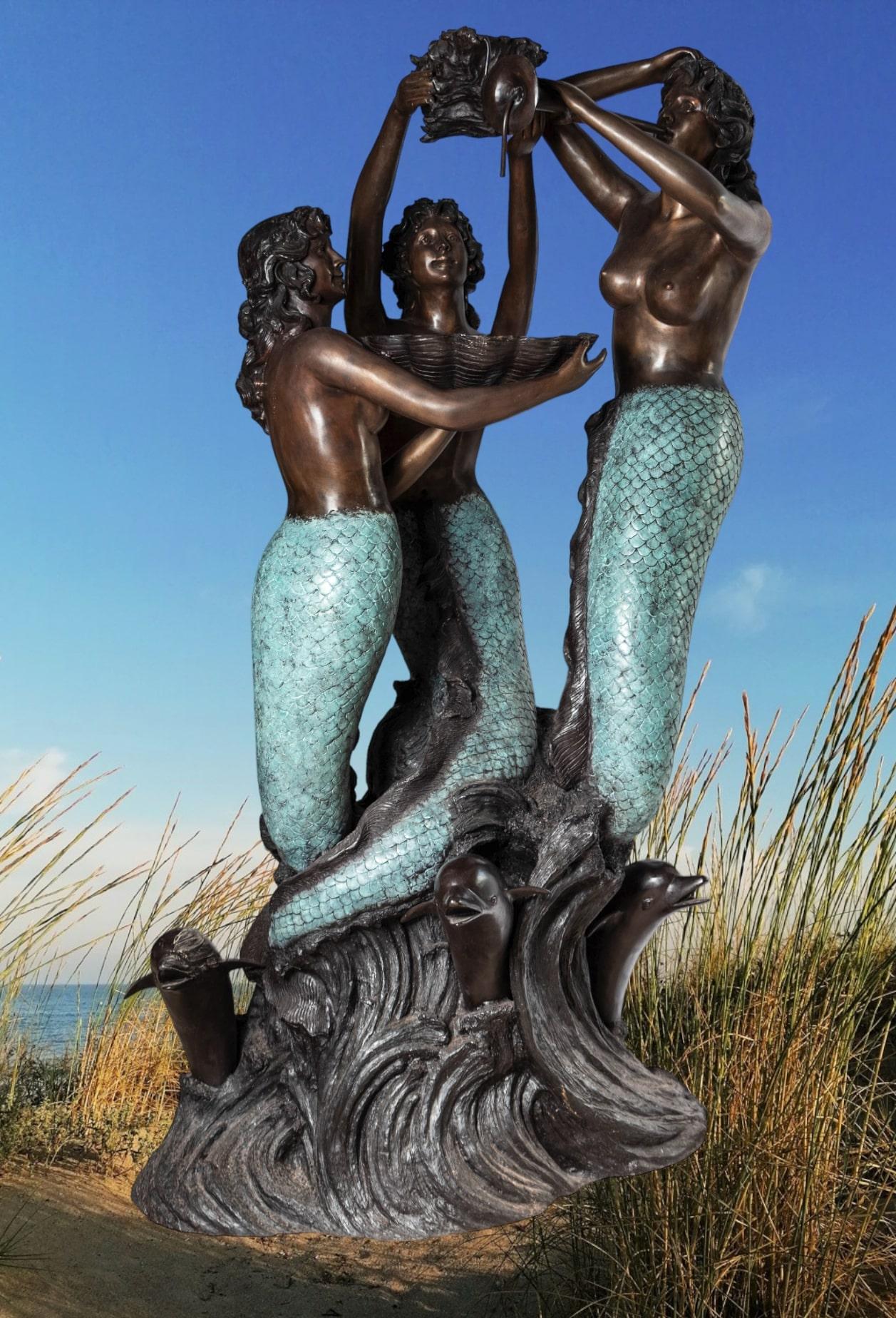 Monumental bronze water fountain depicting three mermaids above the waves surrounded by leaping dolphins. A large shell held by one of the mermaids catches the water from above and returns it to the body of water the statue is (will be) located. In