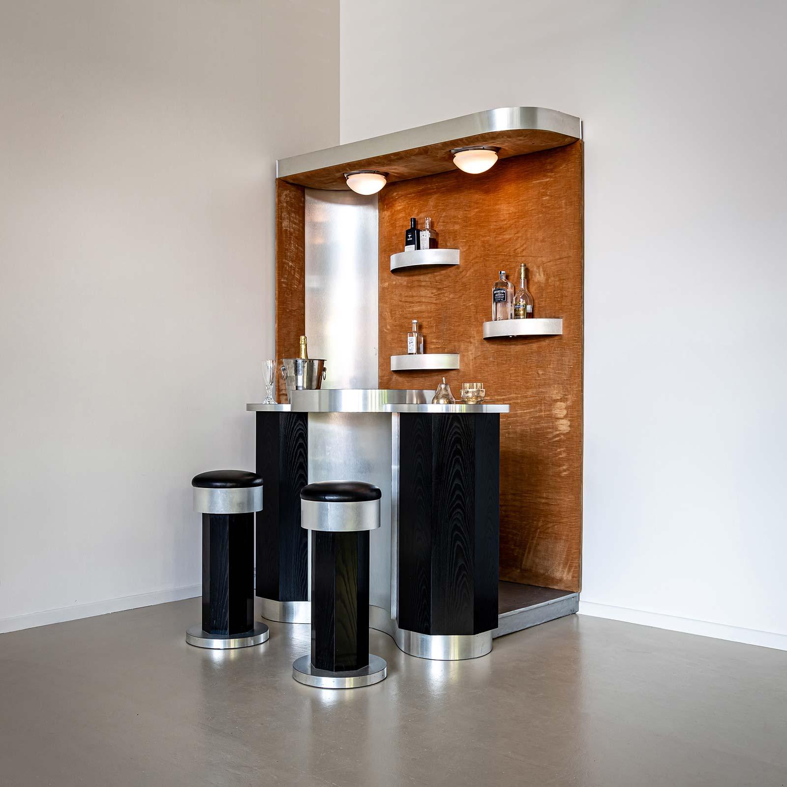 Large corner bar with circular counter and semi-circular shelves on the velvet-lined back wall. The bar is illuminated by two ceiling lamps. Brushed aluminium and black-stained wooden fronts create a high-contrast ensemble. The bar comes with two