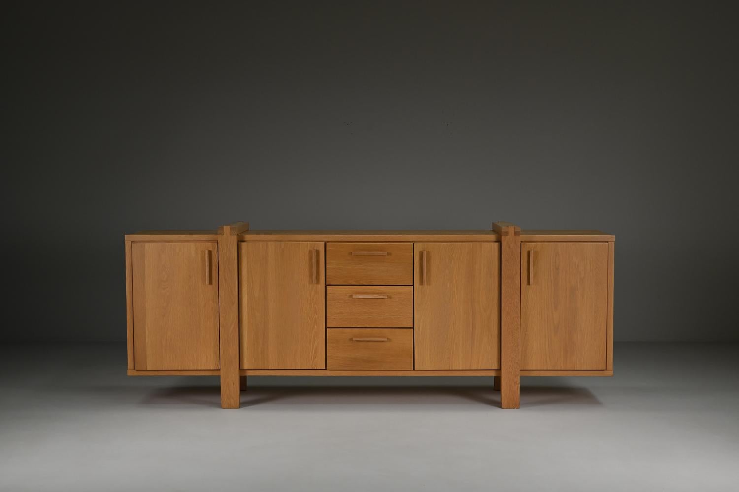 This Belgian sideboard is crafted mostly from solid oak and reflects the Brutalist style with its strong lines and architectural details. The piece offers plenty of storage with four cabinets and three central drawers. Its natural oak finish brings