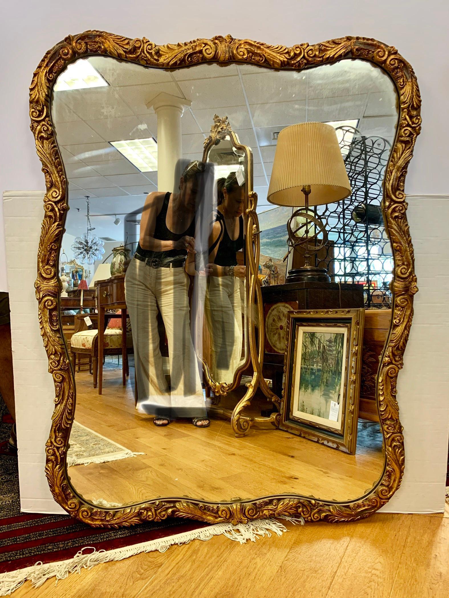 Des courbes élégantes et gracieuses définissent ce miroir en bois doré sculpté. Il présente des volutes sculptées et des motifs feuillus tout autour. Le miroir est prêt à être accroché et dispose de crochets pour les positions verticale et