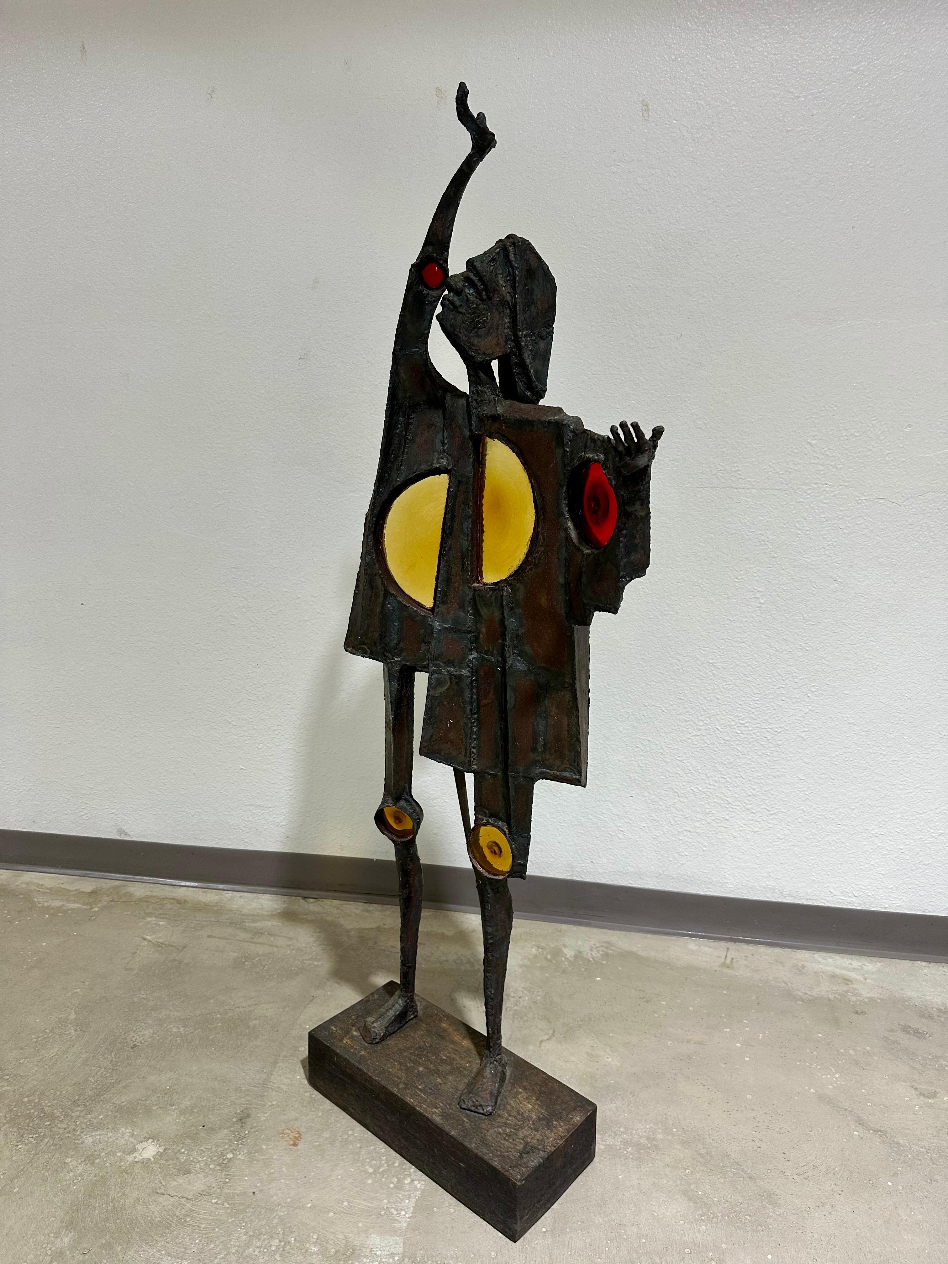 Fantastic large Mid Century Brutalist Figurative Sculpture in Steel with circular glass inserts, on a wood base. Patinated welded steel with red and yellow stained glass depicting a man in a cloak with arms raised. 

Unsigned but well executed,