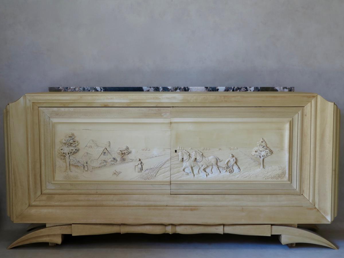 Spectacular and unusual large two-door, blond wood sideboard, featuring intricately sculpted doors. The base has a superb, very graphic line. The doors are elaborately carved in relief, representing an agricultural scene. A slab of grey marble sits