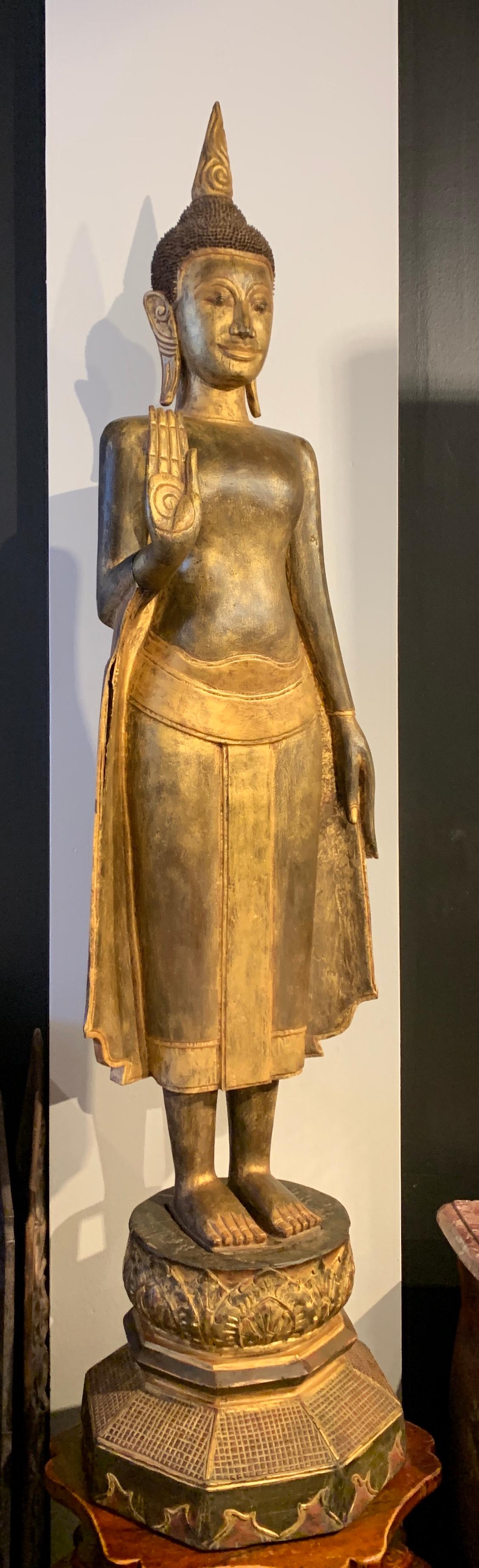 A large, tall Thai standing carved teak wood Buddha, lacquered and gilt, with hand raised in abhaya mudra, far Northern Thailand or possibly Laos, early 20th century. 

Near life-sized and carved from solid teak, the Buddha is portrayed standing