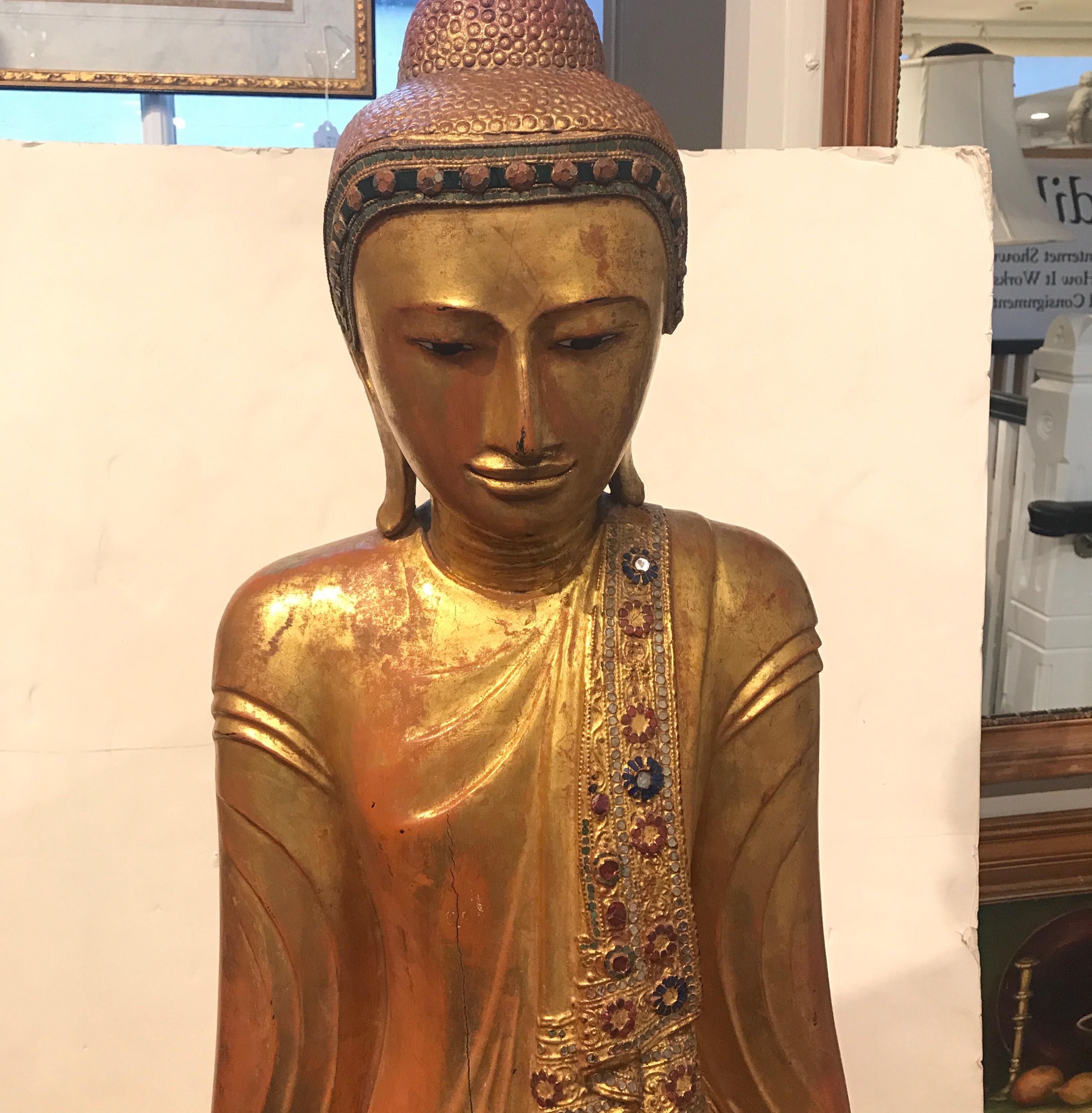 A solid carved wood Thai sculpture of Buddha. The original gold surface with slight wear, decorated with tiny colored mirror trim all around the surface of the clothing. The total height is 61 inches, the total height of just the sculpture is 55
