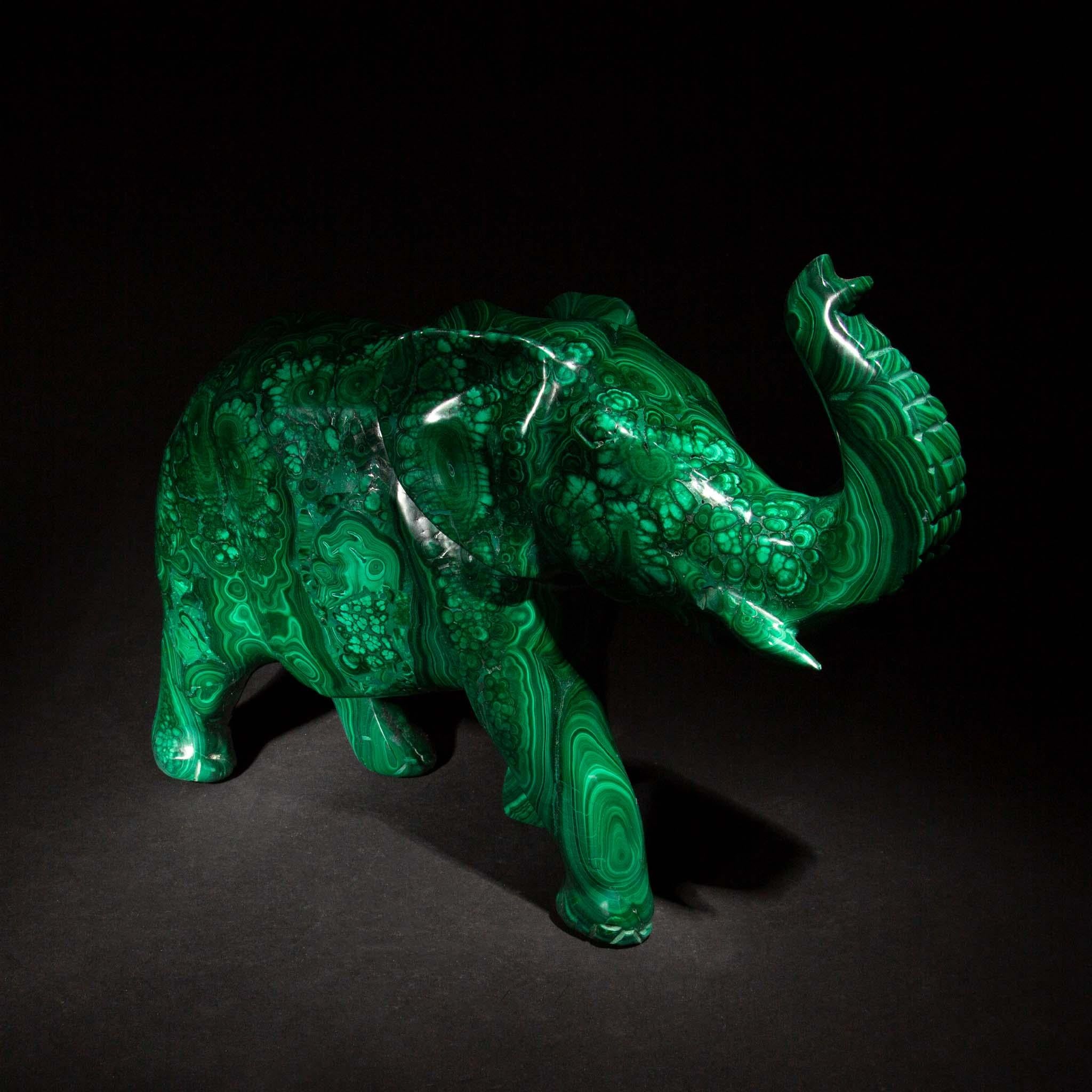This large carved malachite elephant is a stunning display of craftsmanship and the natural beauty of gemstones. Measuring 12 inches in length, 4 inches in width, and standing 9.25 inches tall, it is meticulously sculpted from richly veined