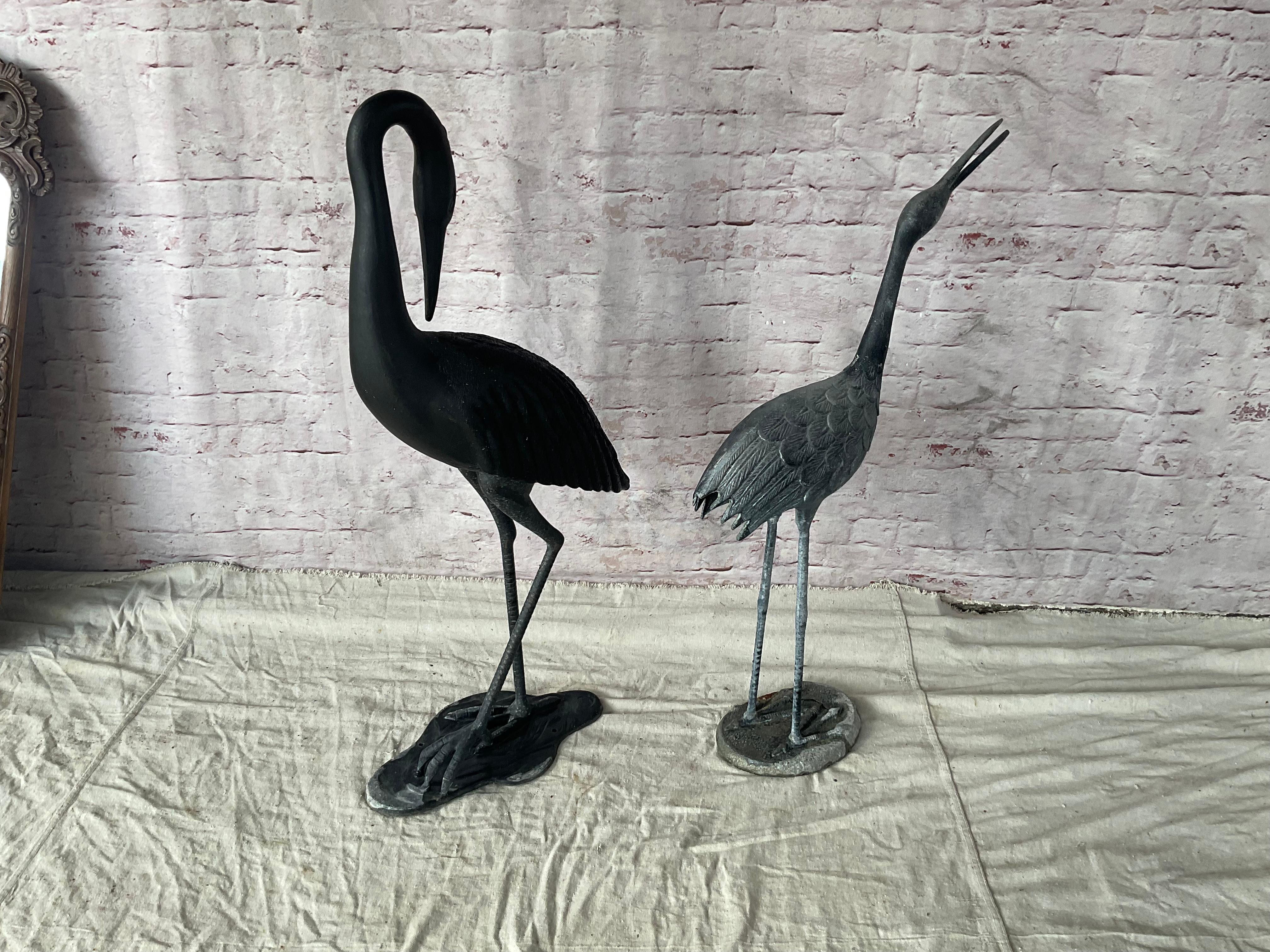 Pair of Cast Metal Crane Sculptures. Cranes are crafted with meticulous attention to detail, capturing the elongated elegance and poised grace of the birds. Standing tall in classic crane poses--one with its neck extended and the other in a more