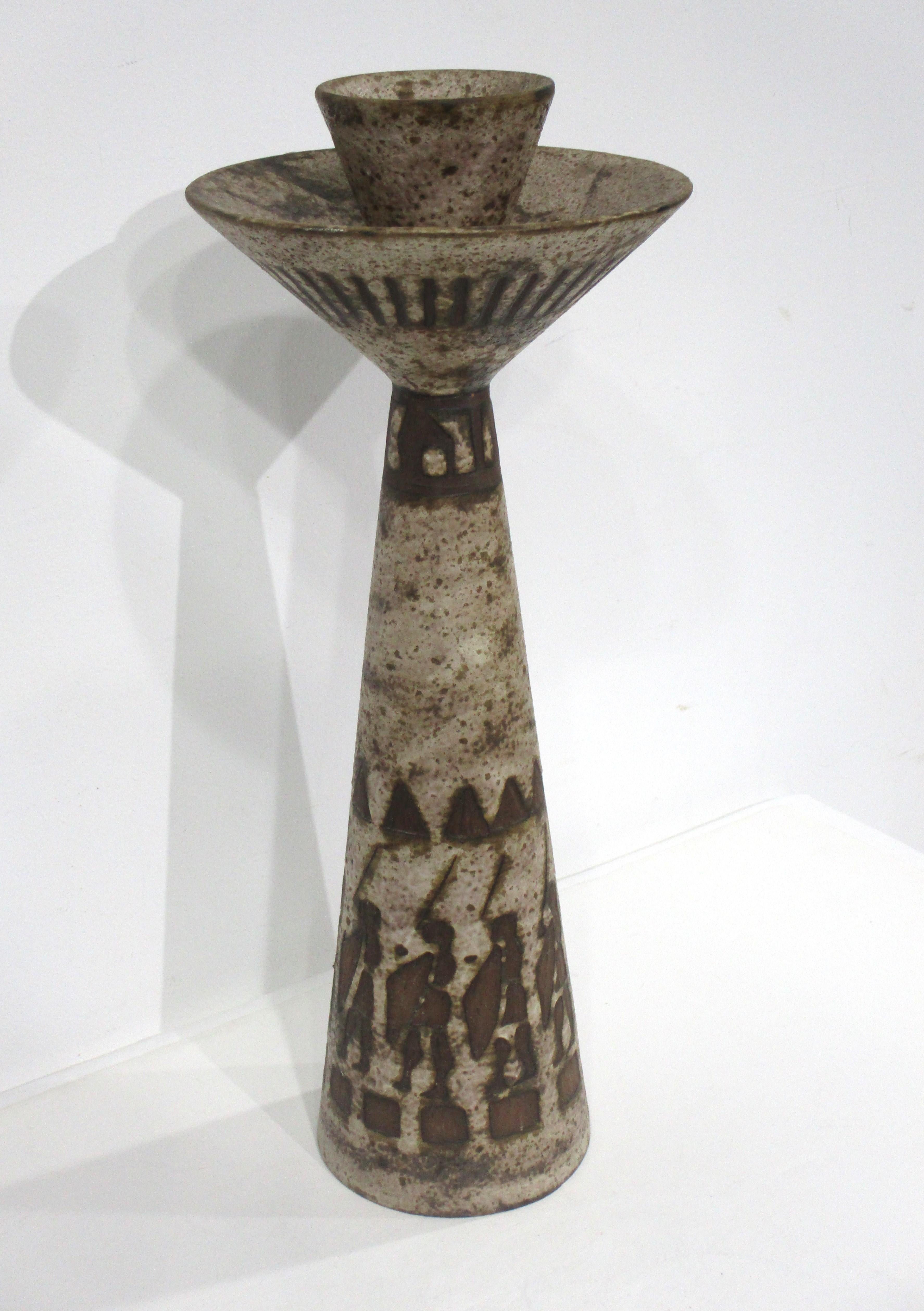 A large sized pottery candlestick with a volcanic styled brown and dark cream glaze . The top has a bigger platform for a larger candle with detailed markings and lower tribal motif to the base . Signed to the bottom by the artist Hans Willing and