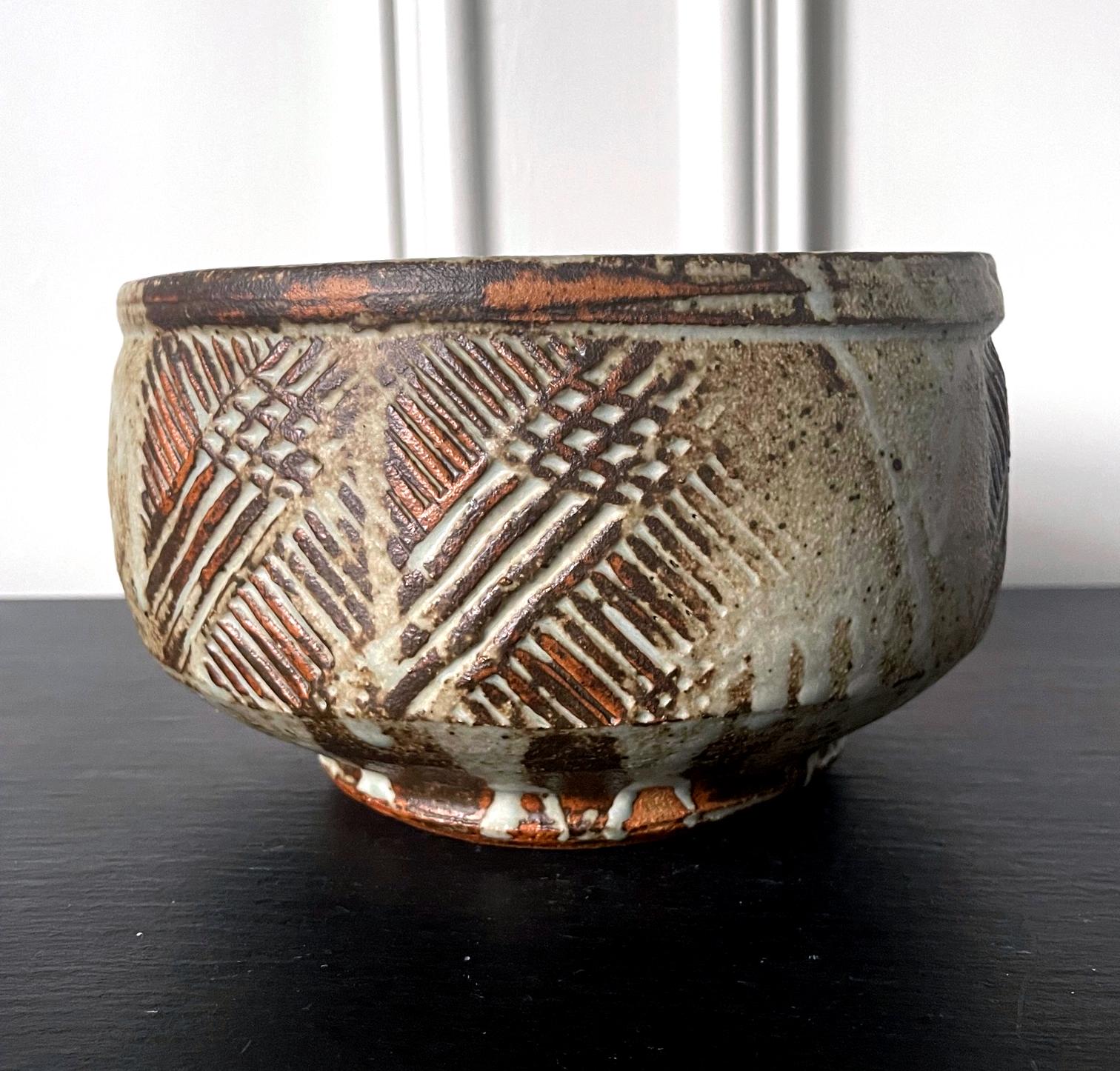 A stoneware center bowl by American ceramist Warren Mackenzie (1924-2018). Thickly potted in an alms bowl shape and supported by a short foot ring, the bowl has a robust and earthy appeal. The surface is craved with hatched lines that cross each